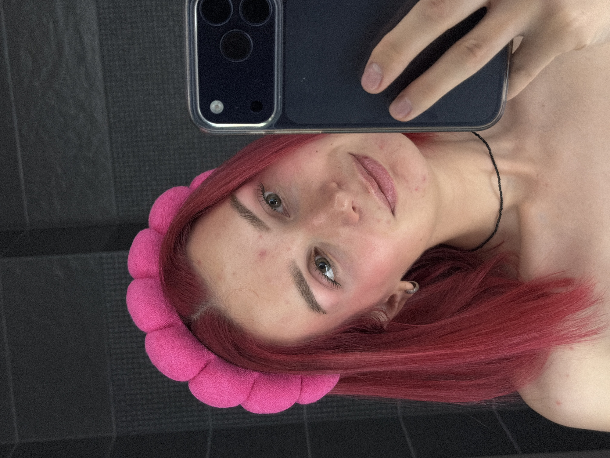 The scene, set in the city of Chaikovsky, Russia, features a young individual with bright fuchsia-red, shoulder-length hair, wearing a thick, padded fuchsia headband with a segmented, bubbly design. The person has fair skin with visible blemishes on the forehead, cheeks, and chin, and wears light pink blush and subtle eye makeup. Their greenish-brown eyes are directed towards the camera. A small silver-colored hoop earring is visible in the left ear, and a thin, dark choker necklace is worn around the neck. The individual's bare shoulders and upper chest are exposed, showing a few small red spots on the right shoulder. A dark-colored smartphone, equipped with a multi-lens camera module and enclosed in a clear case, is held by the person's right hand, partially obscuring the lower right side of their face and neck as they take a mirror selfie. The background consists of a dark, charcoal grey wall with subtle horizontal lines, suggesting an indoor setting.