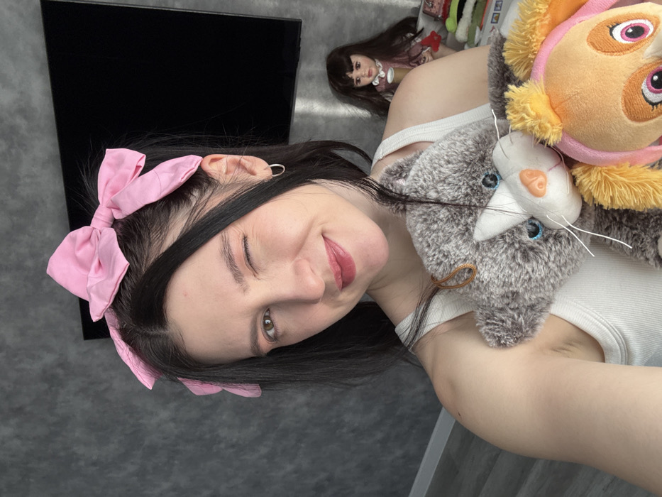 A woman with dark hair and a pink bow winks at the camera. She is holding a gray cat plushie and a Paw Patrol plushie. In the background, there's a black television and a doll. The woman is wearing a white tank top. The background also includes a gray wall.