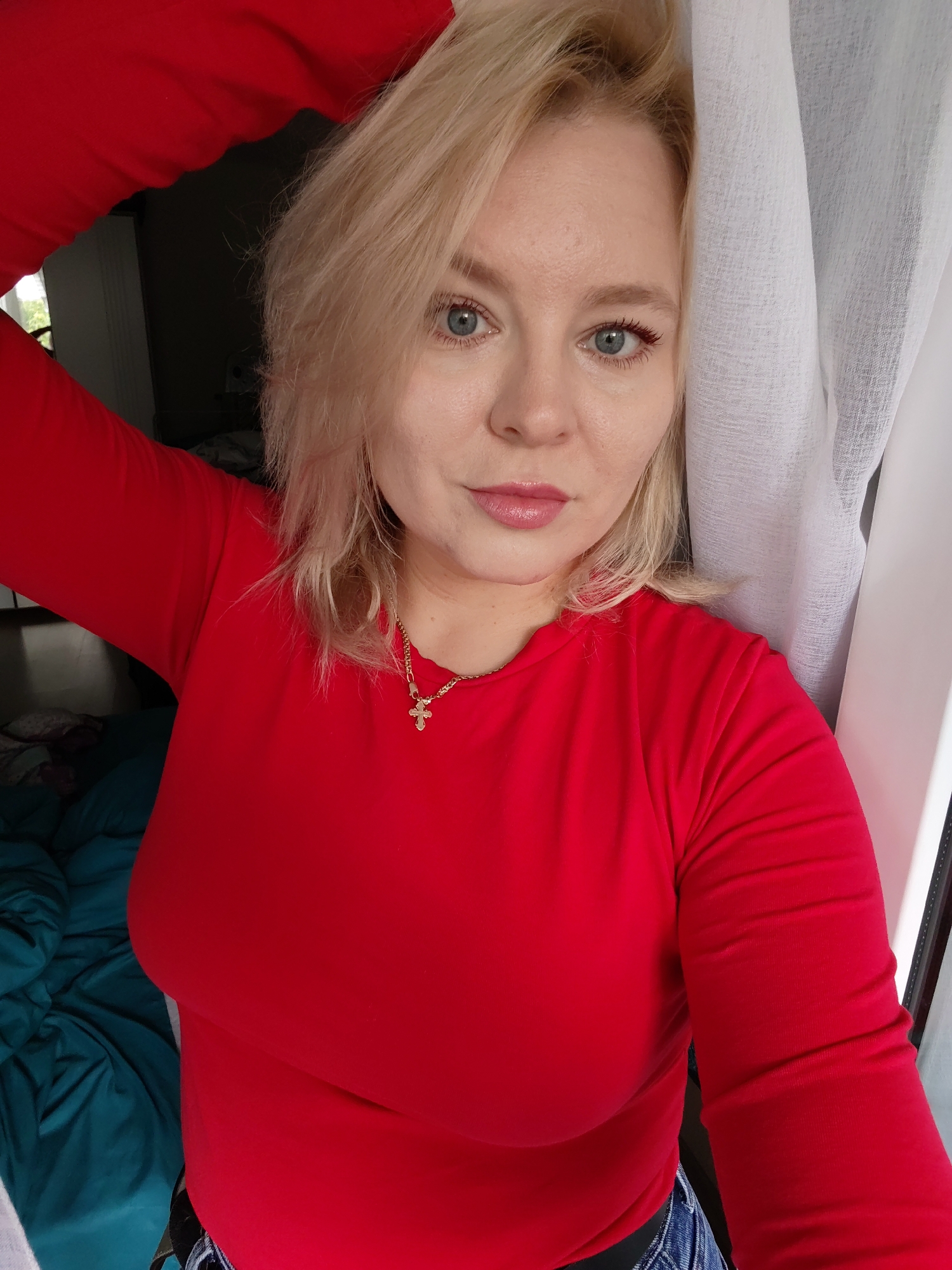 A medium shot image shows a woman with blonde hair and blue eyes, wearing a red long-sleeved shirt and jeans. She is standing next to a white curtain, with a teal-colored duvet visible in the background. The woman is looking towards the camera with her left hand resting on her head. The image appears to have been taken indoors. There is no identifiable information within the image to suggest the location is Izhevsk, Russia.