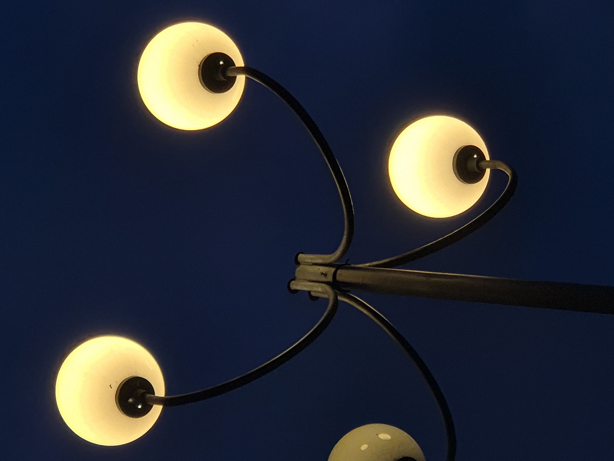 A low-angle shot shows four spherical streetlights affixed to a central metal pole against a dark blue sky. Three of the lights are illuminated, casting a soft, yellow glow. The fourth light, located at the bottom of the frame, is only partially visible and appears unlit, though its surface has some blemishes. The arms connecting the lights to the pole are black and curved. The pole appears to be located in Pavlikeni, Bulgaria.