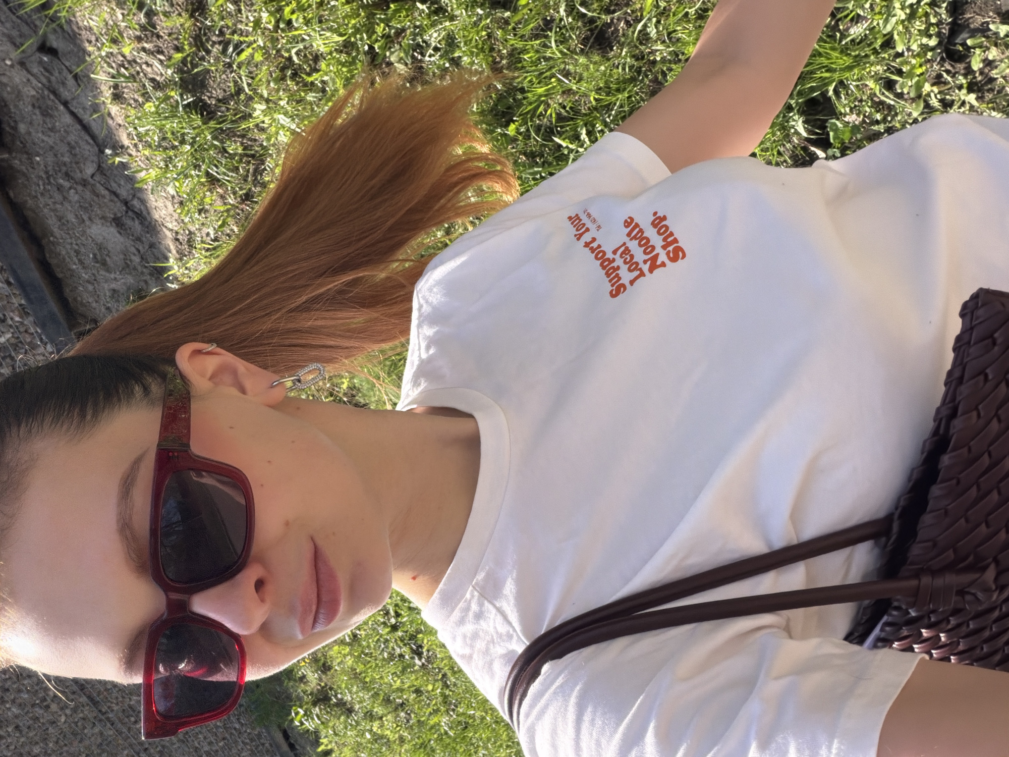 A woman wearing sunglasses and a white t-shirt with the words "Support Your Local Noodle Shop" in orange lettering is shown. She has brown hair pulled back in a ponytail and is wearing earrings. A brown woven bag is slung over her shoulder. She is outside, with grass and a stone pathway visible in the background. There is no indication of the city of Khimki, Russia in the image.