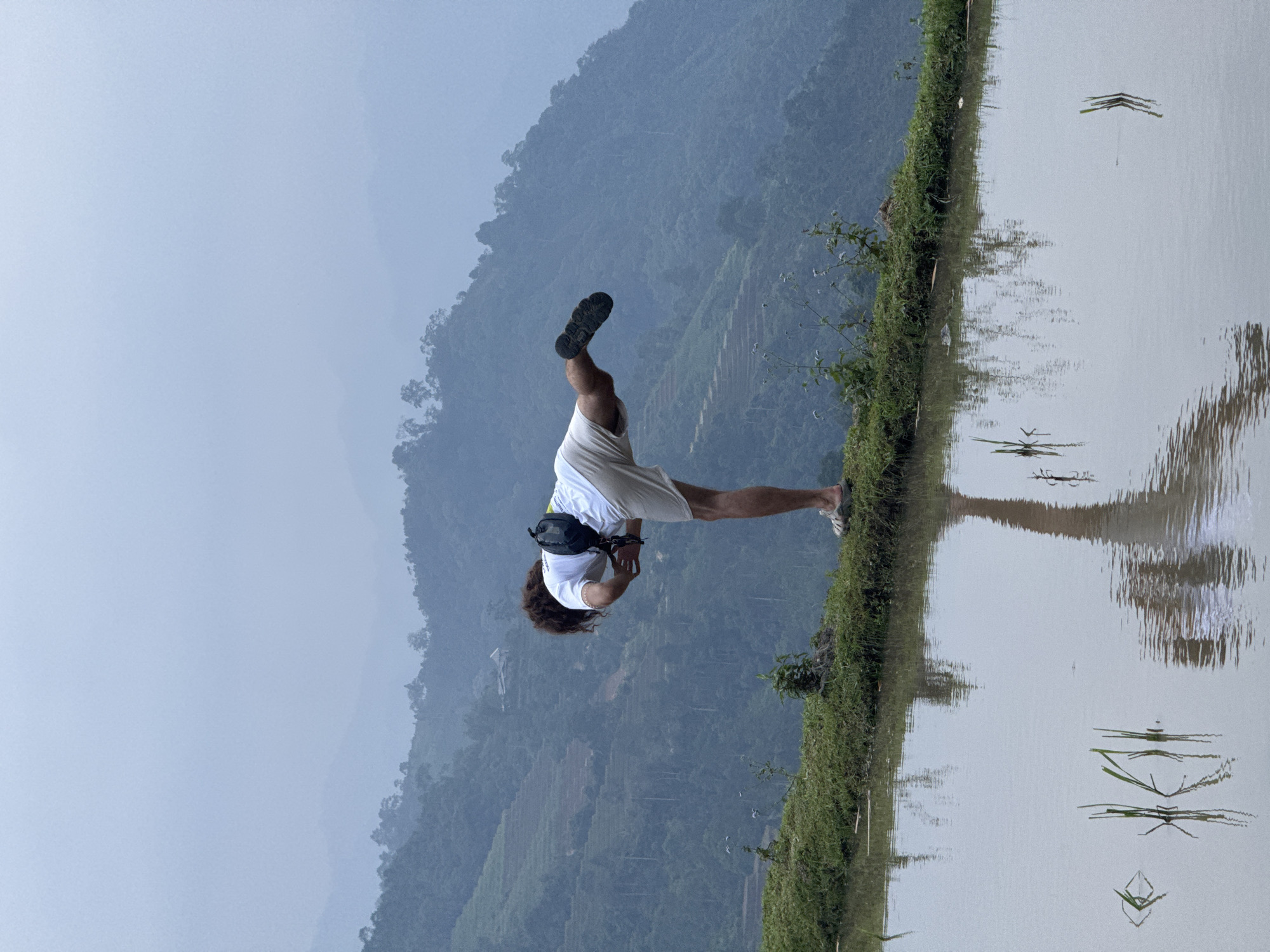 A rotated image captures a person performing an inverted pose, possibly a handstand, on a narrow grassy embankment. The individual, dressed in a white t-shirt, light-colored shorts, and one black athletic shoe, has their head towards the ground and one leg extended upwards, with a black fanny pack visible on their back.
To the right of the person is a still body of water, likely a rice paddy, which highly reflects the overcast sky and elements on the opposite bank, creating a mirror-like effect. Beyond the water and the grassy strip, expansive mountains dominate the background. These mountains are covered in dense greenery and exhibit distinct terraced patterns, indicative of rice cultivation. The mountain peaks are shrouded in a soft haze or mist, blending into a pale, diffused blue-gray sky.
The lighting suggests an overcast day or soft, early morning/late afternoon light, contributing to a serene and somewhat ethereal atmosphere. The landscape cues, including the terraced mountains and rice paddy, strongly place the scene in a rural, agricultural region, consistent with Nậm Dịch Commune, Vietnam. No discernible text is visible in the media.