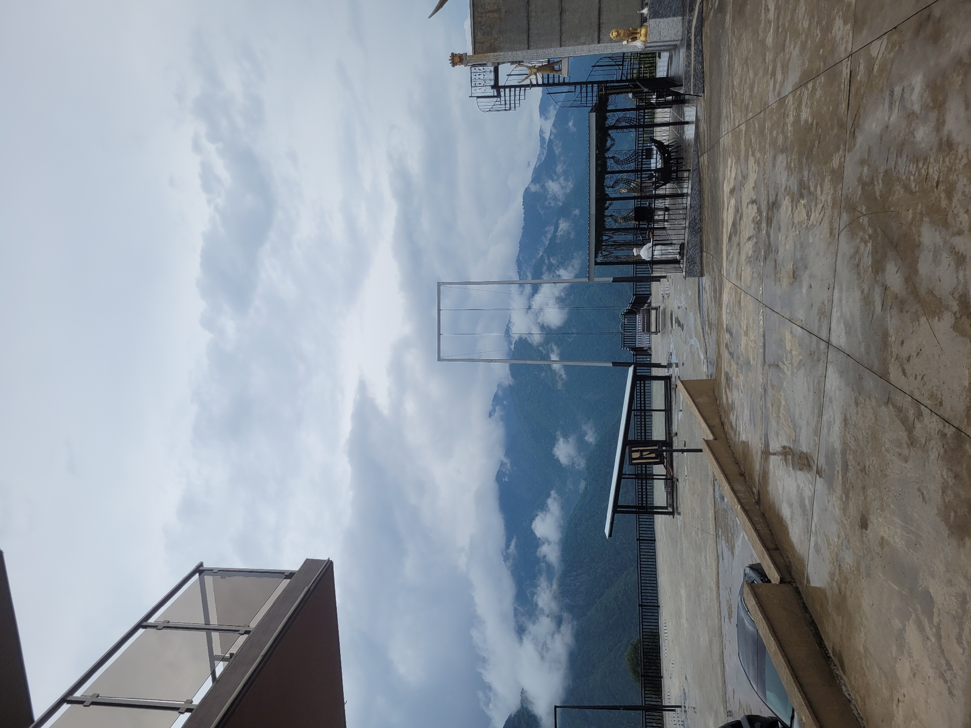 An outdoor paved area with a concrete floor that appears wet and reflective, suggesting recent rain, is shown. In the background, a dramatic mountain landscape shrouded in thick, grey clouds is visible. There is a large, open-air swing set with a tall metal frame in the middle ground. To the right, a spiral staircase leads up to a balcony or platform. Decorative elements such as golden lion statues and figures in white attire are present near the staircase and in a covered seating area. A black car is partially visible on the left side of the frame, parked on the paved area. The environment suggests a scenic, possibly resort-like location in the mountains.