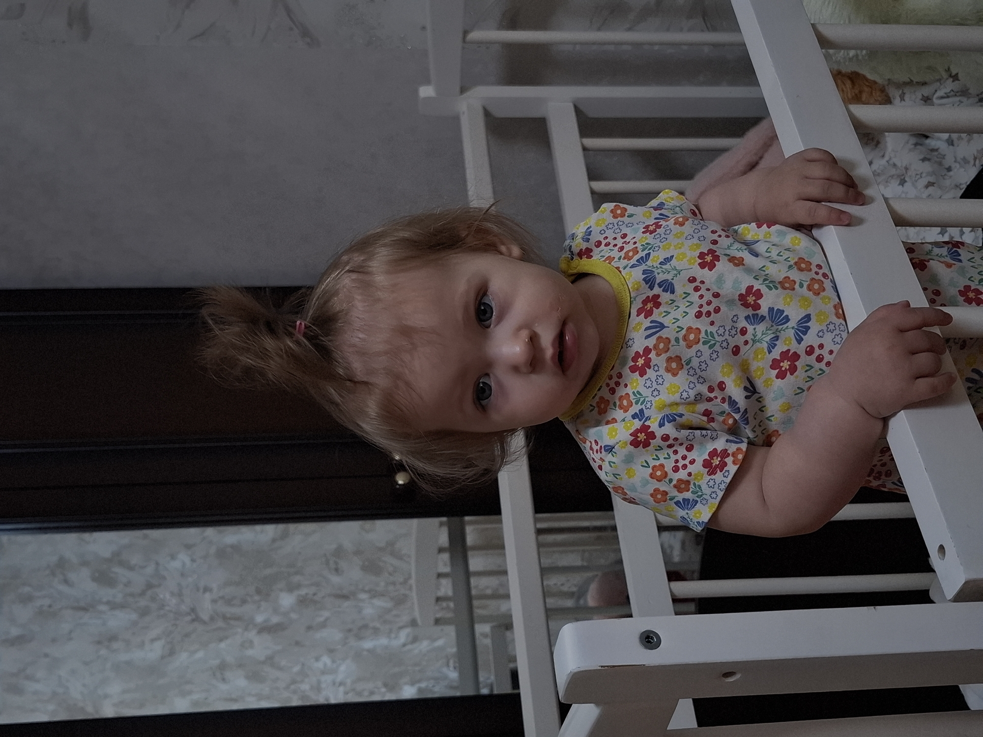 A fair-skinned toddler with light brown hair pulled into a small ponytail is positioned inside a white, wooden baby cot, her hands gripping the side rail. She wears a floral patterned shirt with red, orange, yellow, and blue flowers. The cot appears to be indoors against a wall and near a dark cabinet. The location is Chaikovsky, Russia.
