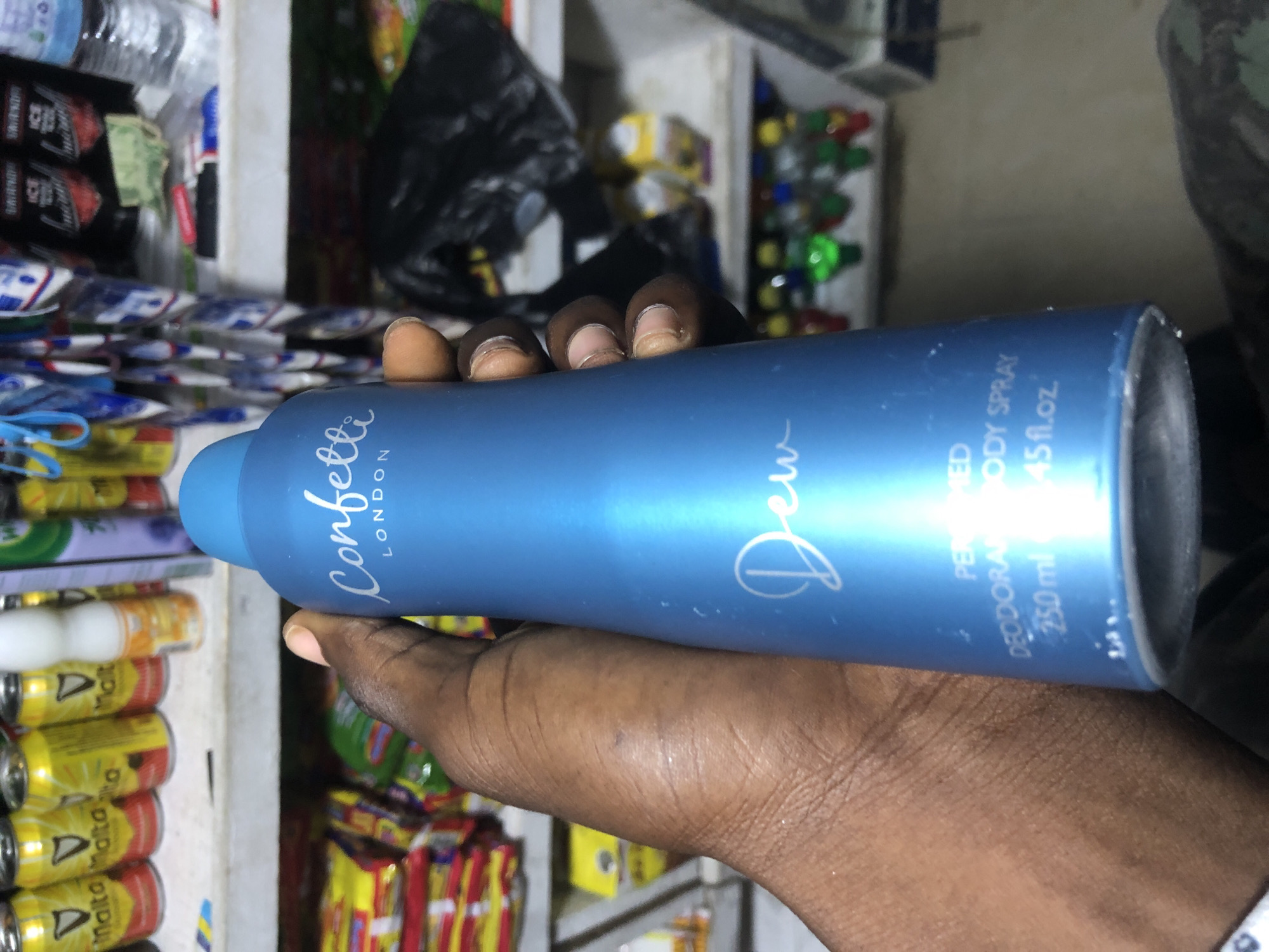 In Ado, Nigeria, a person's hand is holding a blue aerosol can of "Confetti London Dew" perfumed deodorant body spray. The can is decorated with the brand name in white cursive font and "Dew" in a larger white cursive font. Below that, "PERFUMED DEODORANT BODY SPRAY 250 ml 4.45 fl.oz." is printed in white text. The background shows shelves stocked with various goods, including what appear to be cans of drinks and packaged snacks, suggesting a retail environment like a shop or market stall. The lighting is somewhat dim, typical of an indoor setting.