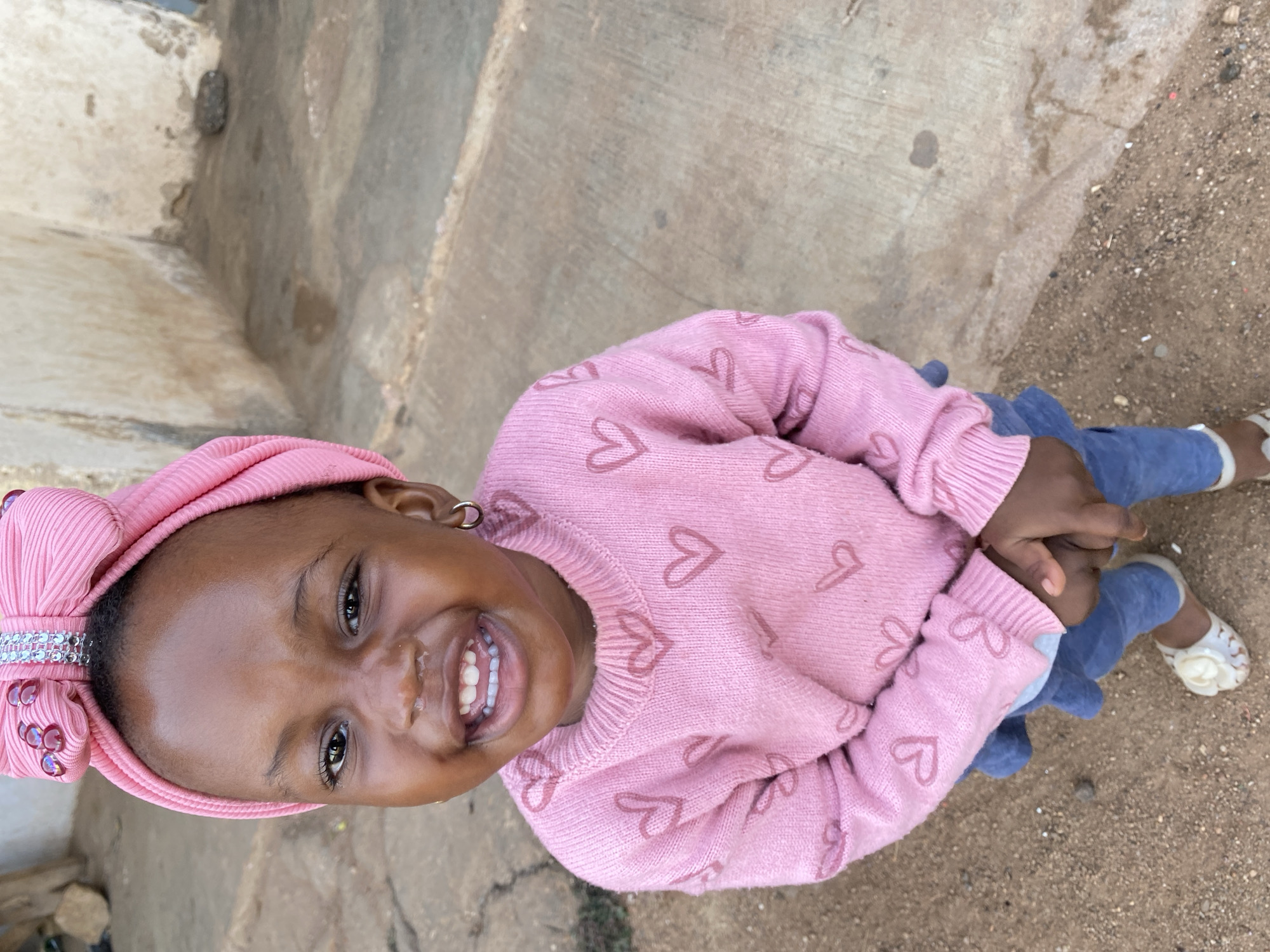 A young African girl stands on a paved surface adjacent to a dirt area in Vom, Nigeria. She wears a pink sweater adorned with hearts, a matching pink headscarf with decorative sequins, and blue pants. White sandals are on her feet. She is smiling, looking directly at the camera. The paved surface shows signs of weathering and discoloration. A wall is visible to the left.