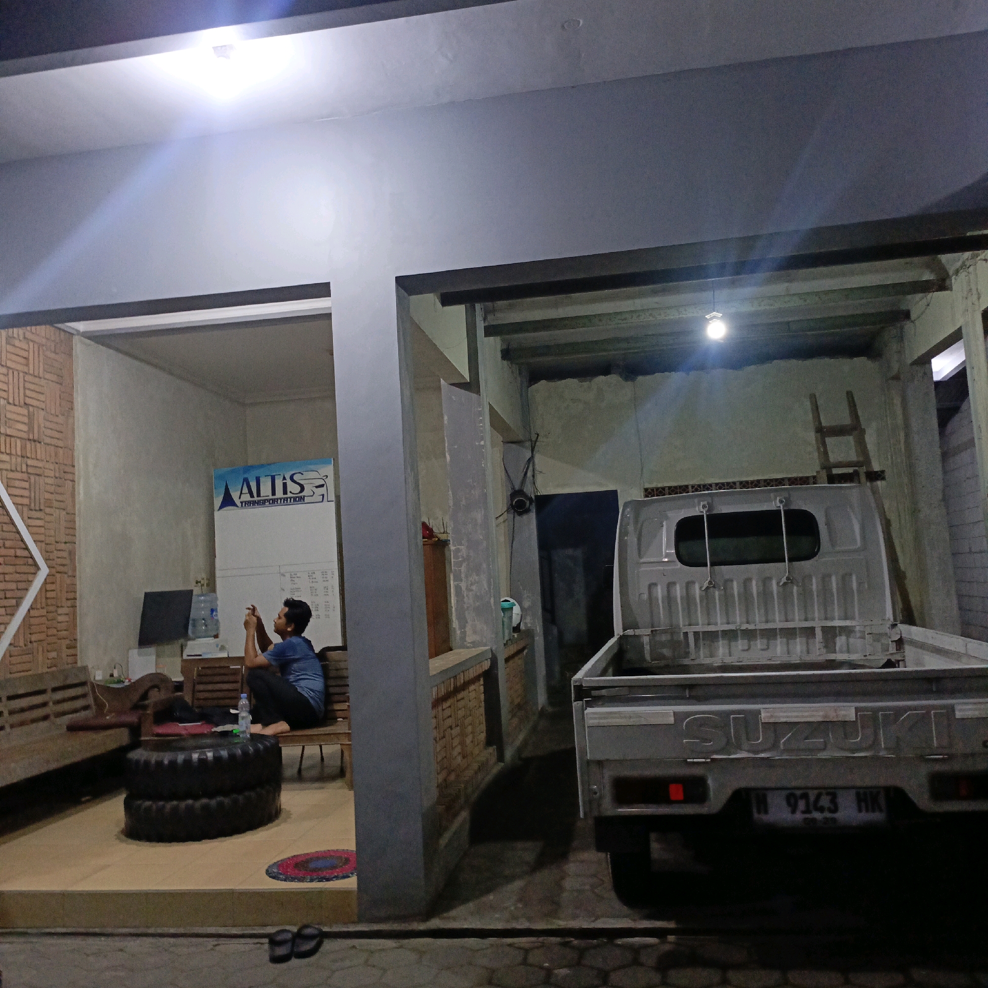 The image depicts an outdoor covered area, possibly a carport or workshop, at night. The scene is illuminated by artificial lighting. In the foreground, a silver Suzuki pickup truck is parked, with its tailgate open. The license plate reads "H 9143 HK". To the left of the truck, a person is sitting on a bench indoors, looking at their phone. They are wearing a blue t-shirt and dark pants. Behind the person is a whiteboard with a logo that appears to say "ALTIS TRANSPORTATION". A water dispenser and a computer monitor are also visible. Two stacked tires serve as a low table in front of the bench. In the background, a wooden ladder leans against a wall. The ground appears to be paved with interlocking bricks. The overall impression is of a functional, utilitarian space. The location is identified as Salatiga, Indonesia.