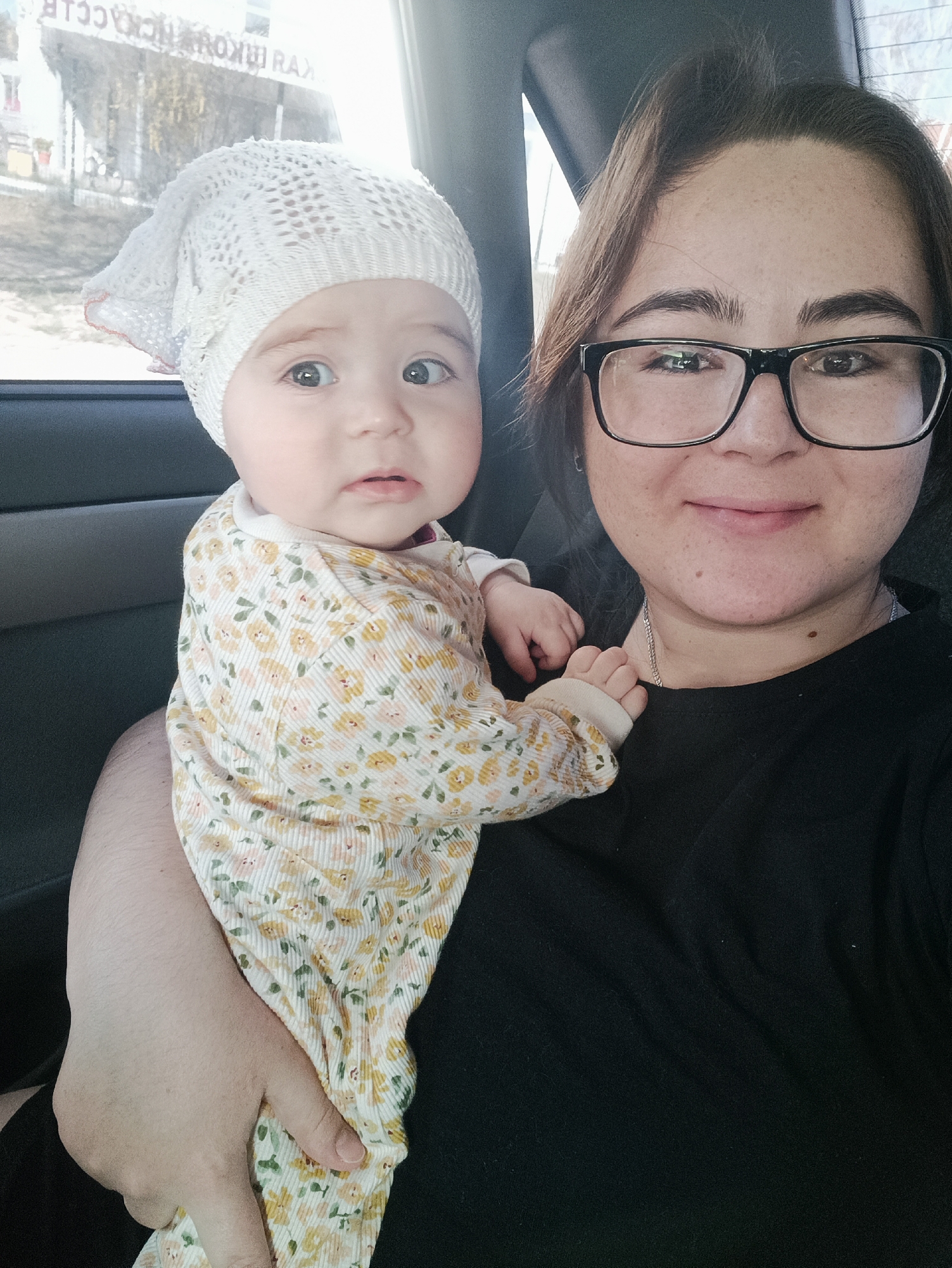 A medium shot shows an adult female with dark brown hair and black-framed glasses holding a baby in her left arm. The baby, wearing a white crocheted hat and a light yellow and white floral patterned onesie, looks towards the camera with wide eyes. The adult female smiles at the camera. They are inside a car, with the interior visible behind them. Reflected in the window behind the baby are structures. Based on the blurred Cyrillic text visible in the reflection, the location is likely Noviy, Russia.