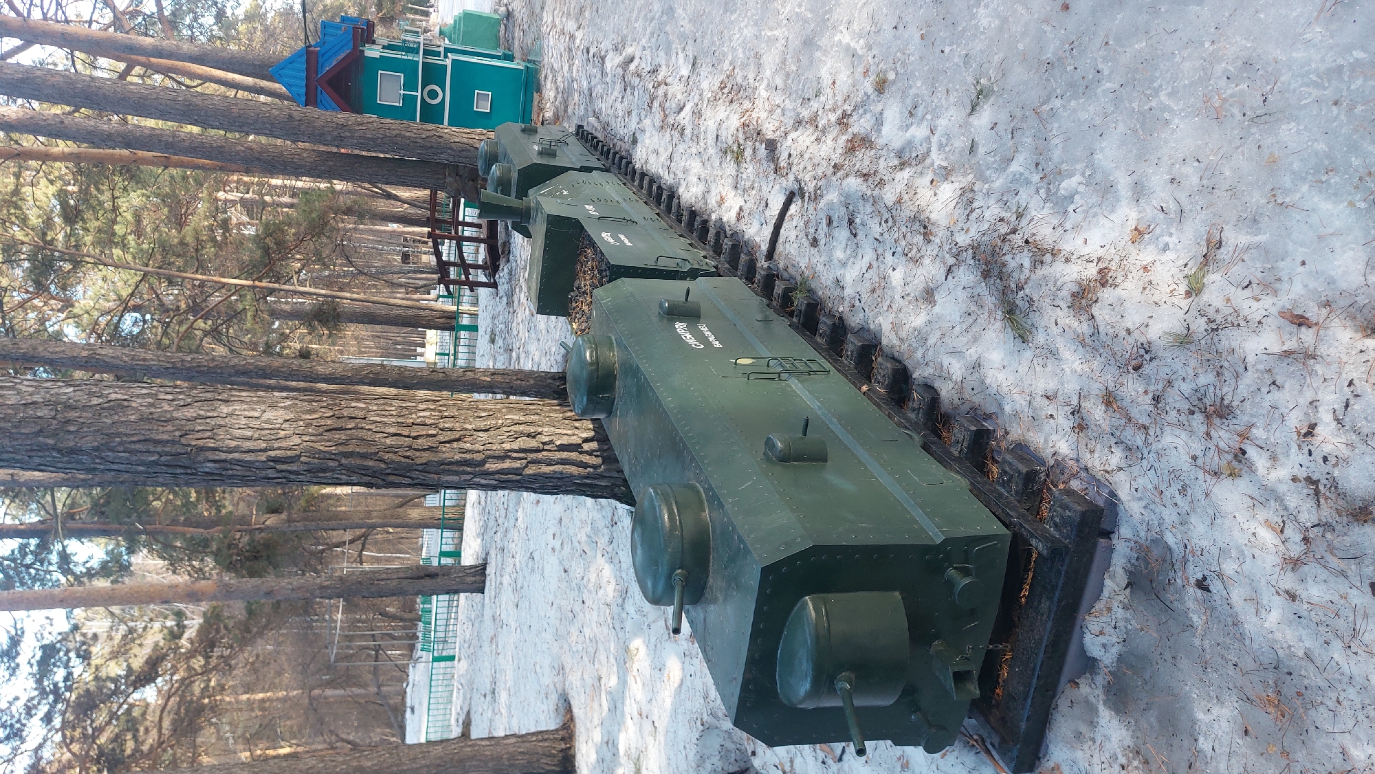 The image shows a model of an armored train in a park-like setting, likely a display piece. The train is painted green and appears to be comprised of several connected sections, each resembling armored vehicles. The train sits on a track amidst a snowy ground cover. Several pine trees are visible in the background, suggesting a wooded area within the park. Further back, a small building with a blue roof and green walls is visible. The location is likely in Novosibirsk, Russia. The scene is static, with no visible people or actions. The overall environment suggests a cold, possibly winter, day.