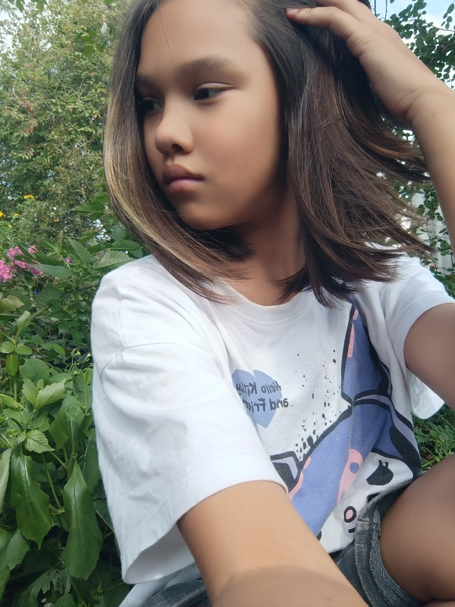 A young girl is captured in a close-up, half-body shot, looking to her left with a pensive expression. She has medium-length, dark brown hair with lighter streaks, which she is touching with her right hand. She is wearing a white t-shirt with a graphic print that includes the text "Hello Kitty" and some abstract shapes in blue, pink, and black. Below the t-shirt, she appears to be wearing denim shorts.
The setting is outdoors, with lush green foliage and some pink flowers visible in the background, suggesting a garden or a natural environment. The lighting indicates it is daytime, with soft sunlight illuminating the scene. The overall mood is relaxed and casual. The location context provided is Karsha, Russia.