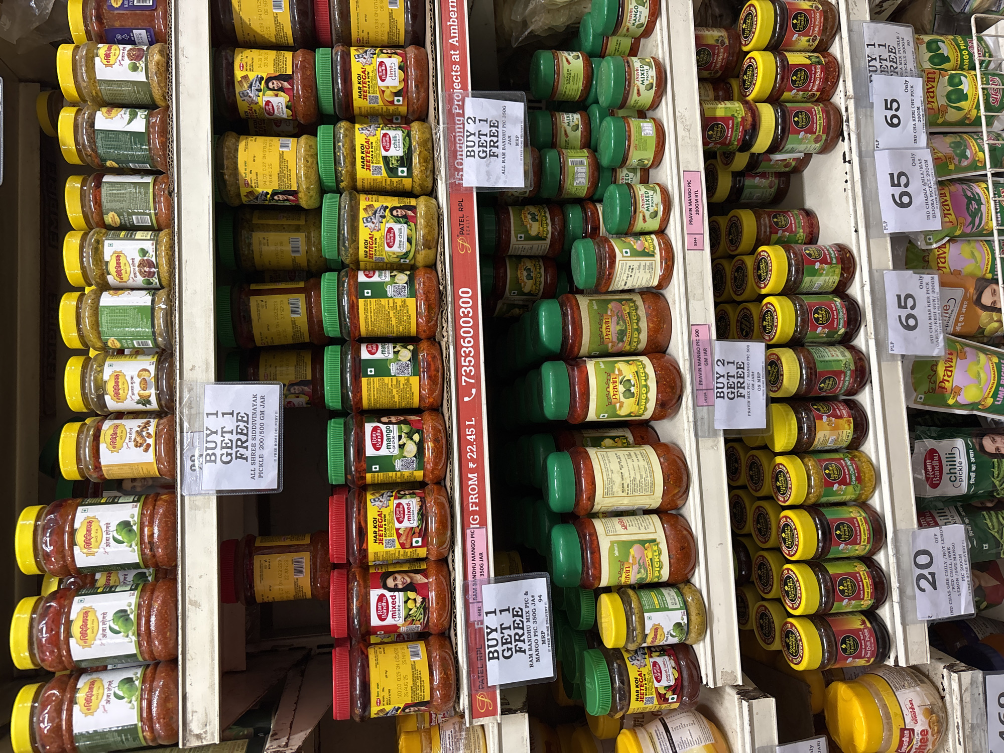 The image shows shelves stocked with various brands and flavors of Indian pickles in jars inside a shop, most likely a grocery store, in Ambernath, India. Several pickle brands, including "Shree Siddhivinayak", "Ram Bandhu," and "Pravin," are visible. The jars are of different sizes and colors, with labels indicating the type of pickle. Price tags and promotional offers, such as "Buy 1 Get 1 Free" and "Buy 2 Get 1 Free," are displayed. The environment appears to be a typical retail setting.