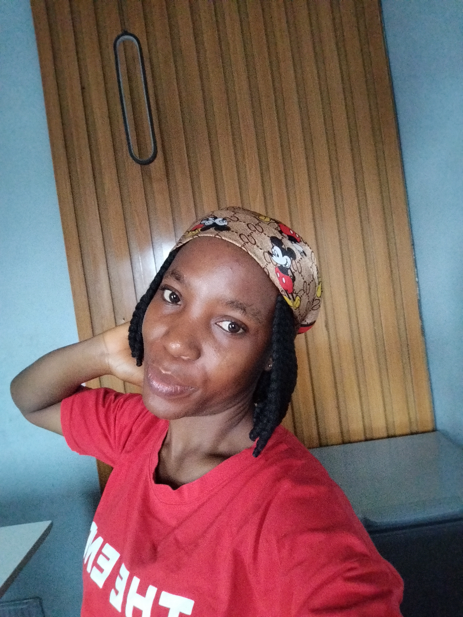 A young woman with dark skin and dark hair braided into two pigtails is captured in a close-up selfie taken from a high angle. She is wearing a red t-shirt with white lettering that appears to be reversed and a headscarf patterned with Mickey Mouse. Her left arm is raised and her hand is behind her neck. She is looking directly at the camera with a slight smile.
The background consists of a light blue wall on the left and a wooden panel wall on the right. The wooden wall has vertical grooves and an oval-shaped, dark-lined indentation in the upper center. A dark grey couch or bench is visible in the lower right corner of the frame. The lighting suggests an indoor setting. The image does not contain specific landmarks or contextual clues that would allow for precise identification of the location within Iguosa, Nigeria.