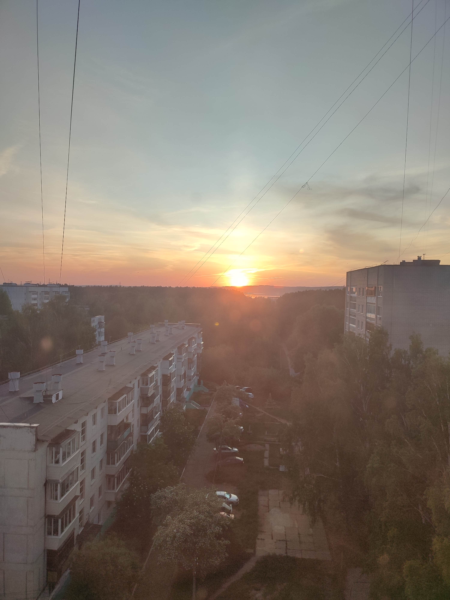 The image shows a cityscape at sunset. Two large, multi-story apartment buildings dominate the foreground, with rows of balconies and windows. Trees surround and partially obscure the buildings. In the distance, the sun sets over a horizon of trees and possibly a body of water. Overhead power lines crisscross the sky. The overall tone is warm due to the sunset.