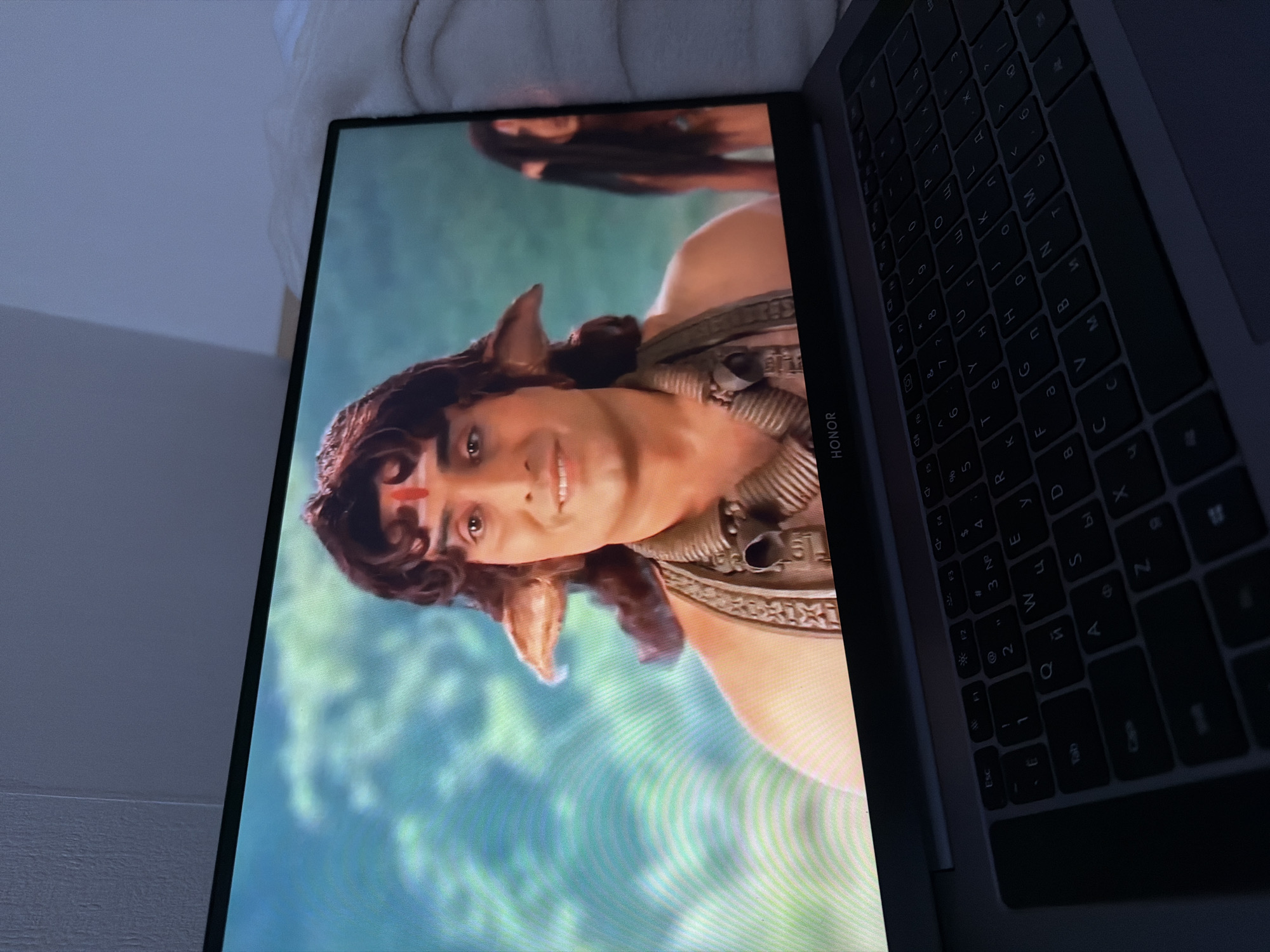 Here's a detailed description of the image:
The image shows a close-up of a laptop screen displaying a scene with a man who appears to be of South Asian descent. He has brown, curly hair, light skin, and is wearing what seems to be ancient or mythological attire, including ornate necklaces and shoulder pieces. A red mark is visible on his forehead. Additionally, he has pointed, elf-like ears. The background appears to be a light blue or green abstract pattern, possibly suggesting water or sky.
The laptop keyboard is partially visible in the foreground, showing dark keys with white lettering. The brand "HONOR" is printed on the hinge area of the laptop.
There's no indication of a location in Saint Petersburg, Russia, or any information to identify this image as being from there.