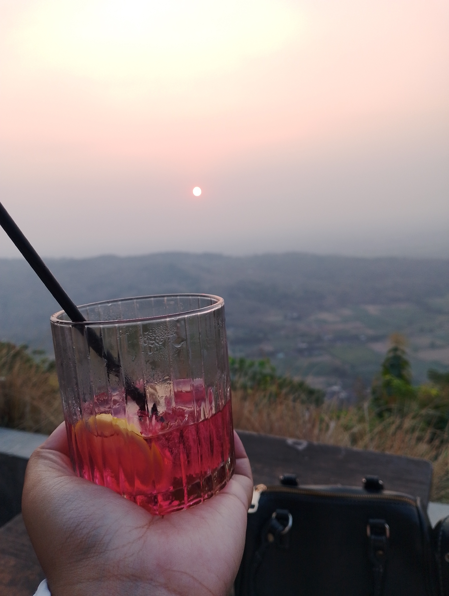 A person's hand holds a glass of pink liquid with ice and a lemon slice, and a black straw. The glass is positioned in the foreground, with condensation on its exterior. In the background, a hazy sunset paints the sky in soft pink and orange hues, with the sun visible as a small, bright orb. Beyond the sky, rolling hills stretch into the distance, their details softened by atmospheric perspective. A black handbag rests on a wooden surface in the middle ground. The scene appears to be set at an elevated viewpoint, possibly a restaurant or cafe, overlooking a landscape.