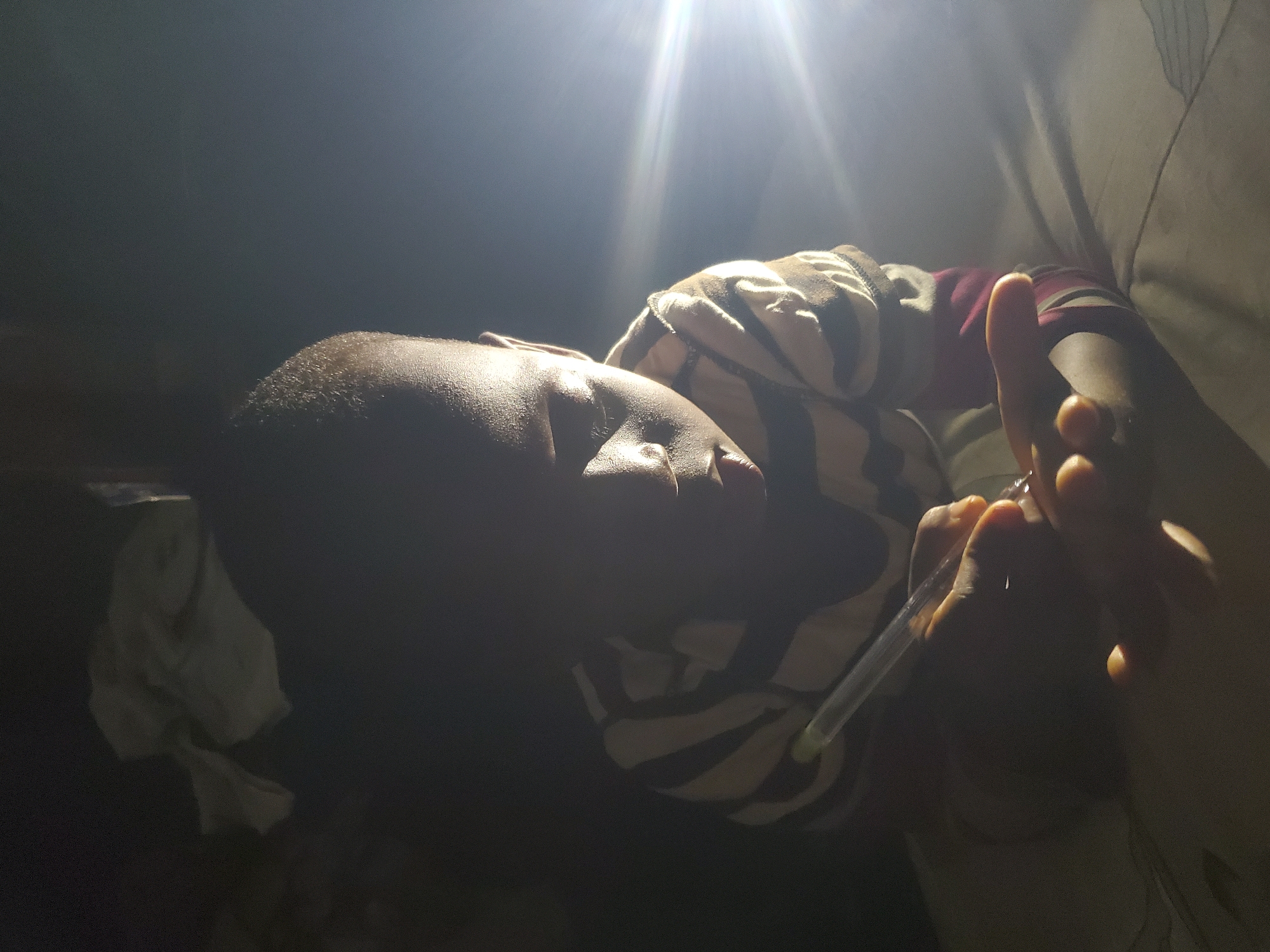 A person lies in a dimly lit area, possibly indoors given the presence of what appears to be a cloth or fabric backdrop. The person is holding a thermometer. The location is Unknown, Unknown.
