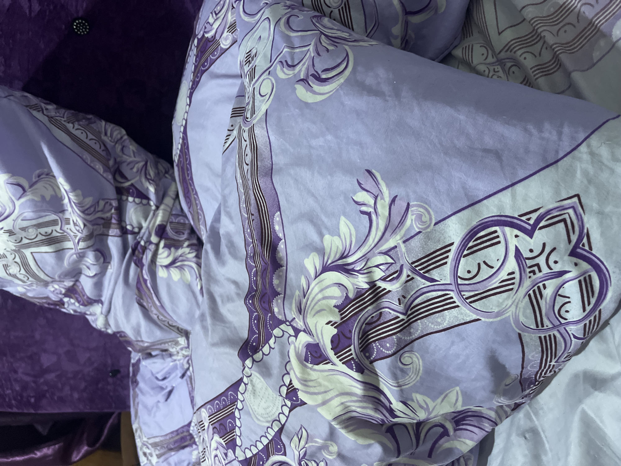 This close-up image captures a section of bedding and an adjacent piece of furniture, likely within a bedroom setting. The primary focus is on a duvet or blanket characterized by an ornate pattern. This intricate design features stylized floral motifs, flowing scrolls, and geometric lines rendered in varying shades of purple (lavender to deep violet) and white or light cream. The fabric appears to be smooth, possibly cotton or a synthetic blend, showing numerous folds and creases.
To the left of the bedding, a dark purple, plush material is visible, which seems to be part of a headboard or upholstered furniture. This material exhibits a velvet-like texture and includes a single, dark, button-like embellishment. A plain white or off-white sheet or blanket can be seen along the right edge and bottom right corner of the frame, providing a contrasting background to the patterned duvet.
No people are present in the image, and no specific activity is depicted, though the scene clearly indicates a sleeping area. The lighting appears to be indoor and artificial, highlighting the textures and patterns of the fabrics. There is no visible text in the image. The context provided indicates this scene is located in Abuja, Nigeria.