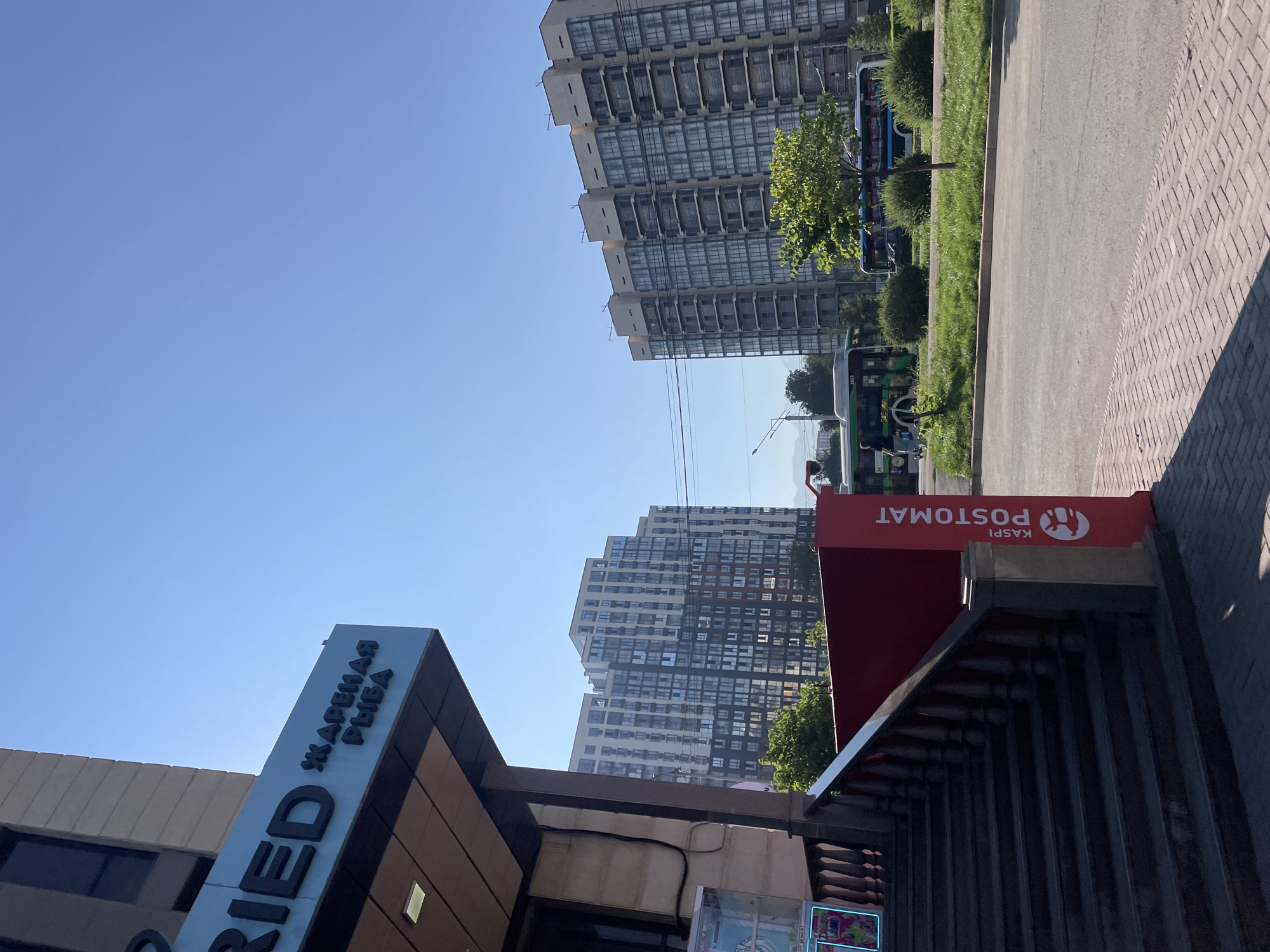 This is a street-level, eye-level, medium shot of a commercial building with apartments above, and a street with traffic in front, taken during the day in Almaty, Kazakhstan. The building on the left has signage that reads "RIED" and "ЖАРЕНАЯ РЫБА" in black lettering on a blue background. In front of the building, a red sign with white lettering reads "KASPI POSTOMAT". To the right of the building, there are two tall apartment buildings with many windows. A street runs in front of the apartment buildings, with two buses and some greenery. The sky is clear and blue.