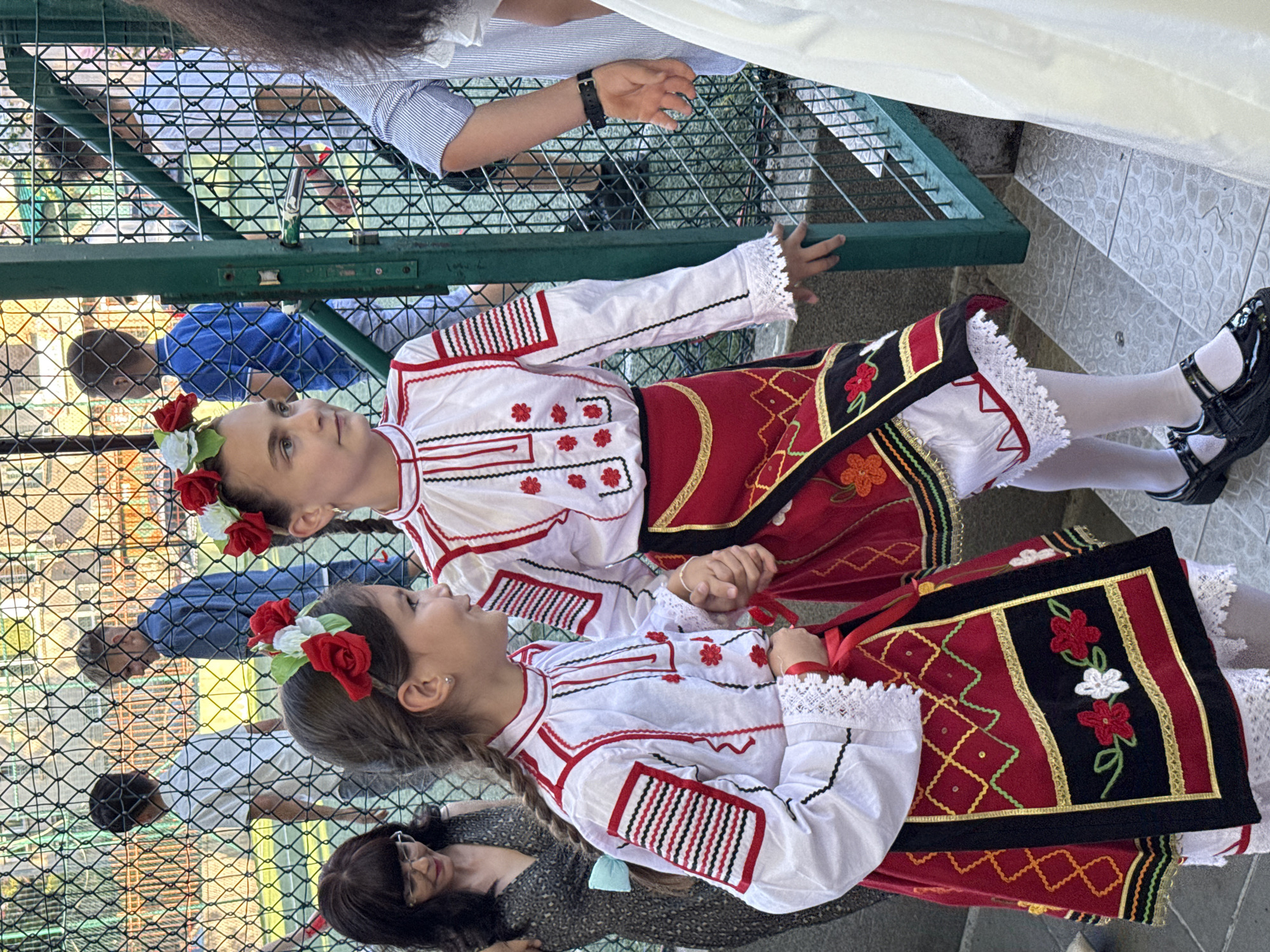 Two young girls in traditional Bulgarian folk costumes are walking down a flight of outdoor stairs, with a metal fence to their left and a green metal gate to their right. The girl in the foreground on the left wears a white blouse with red and black embroidery on the sleeves and chest, and a red skirt with a black embroidered panel. Her hair is braided and adorned with red and white flowers. She is holding hands with the girl next to her. The second girl, partially visible, also wears a similar folk costume. Behind the fence, other children and adults are visible, some on what appears to be a sports field. The scene appears to be a public space in the city of Pleven, Bulgaria, possibly during a cultural event or festival.