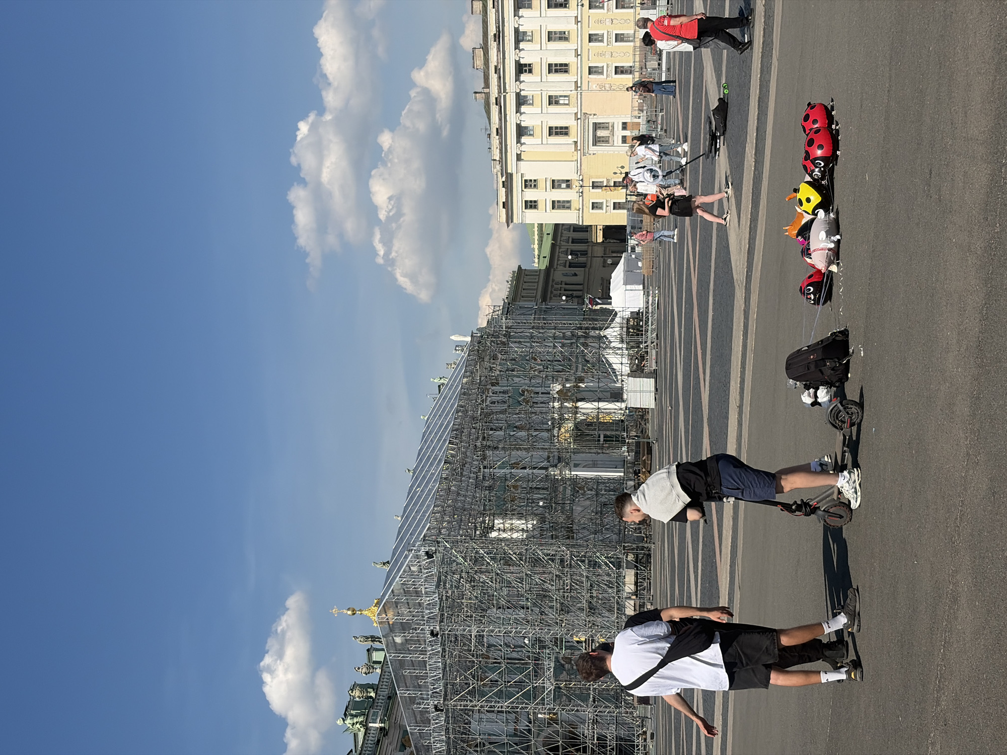 The image captures a sunny day in Palace Square, Saint Petersburg, Russia. In the foreground, two young men stand in the plaza, one on a scooter. Nearby, several colorful balloons shaped like ladybugs and other figures are laid out on the ground, possibly for sale. In the mid-ground, a group of people are gathered, some appearing to be a film crew with equipment. Further back, a large building is partially covered in scaffolding, suggesting ongoing construction or renovation. The background features the ornate architecture characteristic of Saint Petersburg, with classical buildings and a clear blue sky dotted with white clouds.