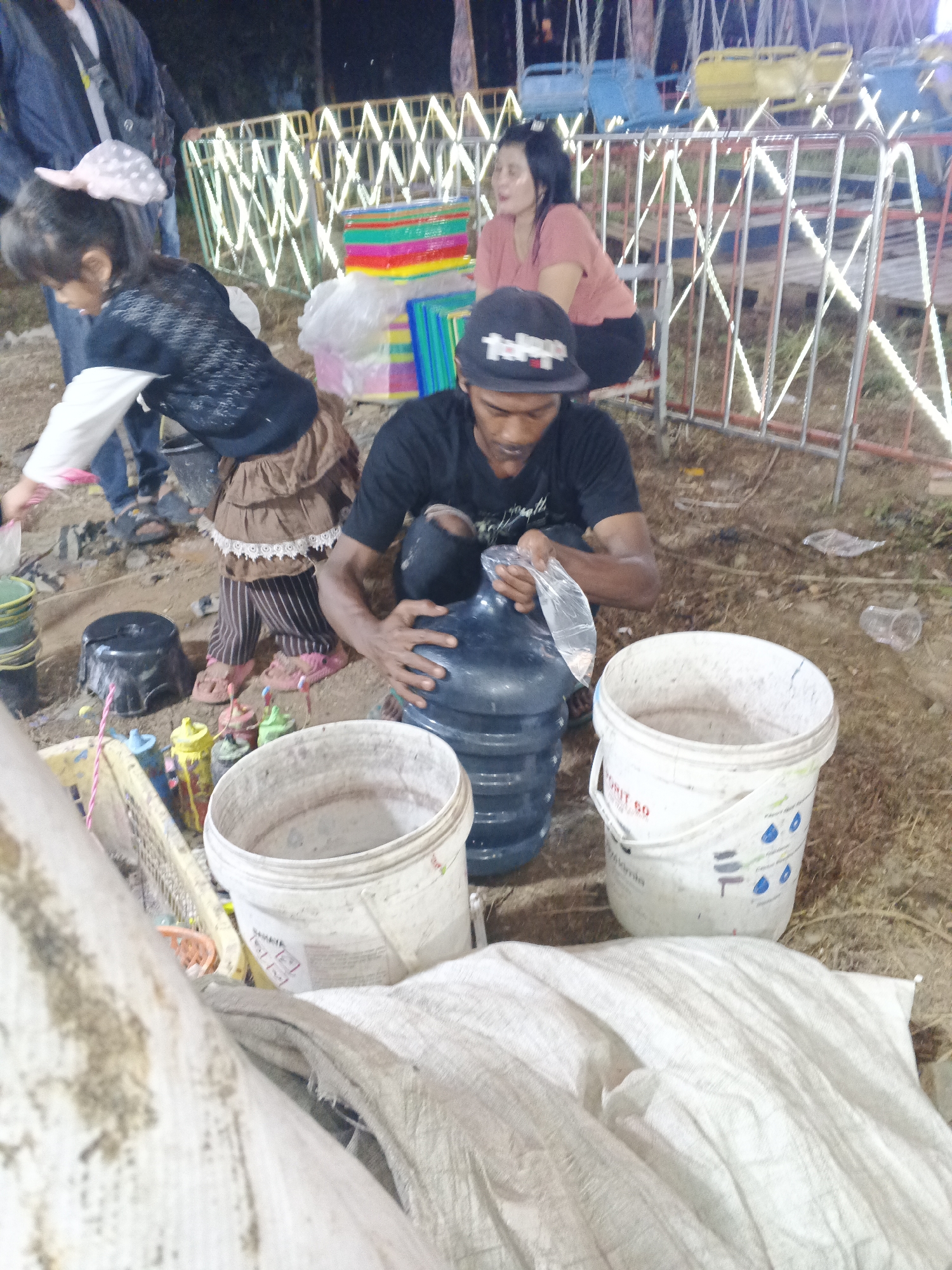 In Sukabumi, Indonesia, at an outdoor event likely a carnival or festival, a man is seen filling a large plastic water jug with a clear plastic bag. He is wearing a black t-shirt and a black baseball cap. Beside him are two large white buckets, one with text and graphics indicating it's a paint bucket, and a stack of colorful plastic containers. A young girl with a large pink bow in her hair and wearing a brown skirt is standing nearby, also appearing to be involved in the activity. She is wearing a black and white patterned top and striped pants. In the background, a woman in a pink shirt sits behind a metal fence adorned with bright white lights, likely part of a ride. The ground is dirt, and there are other people and objects scattered around, suggesting a bustling environment. The overall scene depicts a moment of activity within a lively outdoor setting.