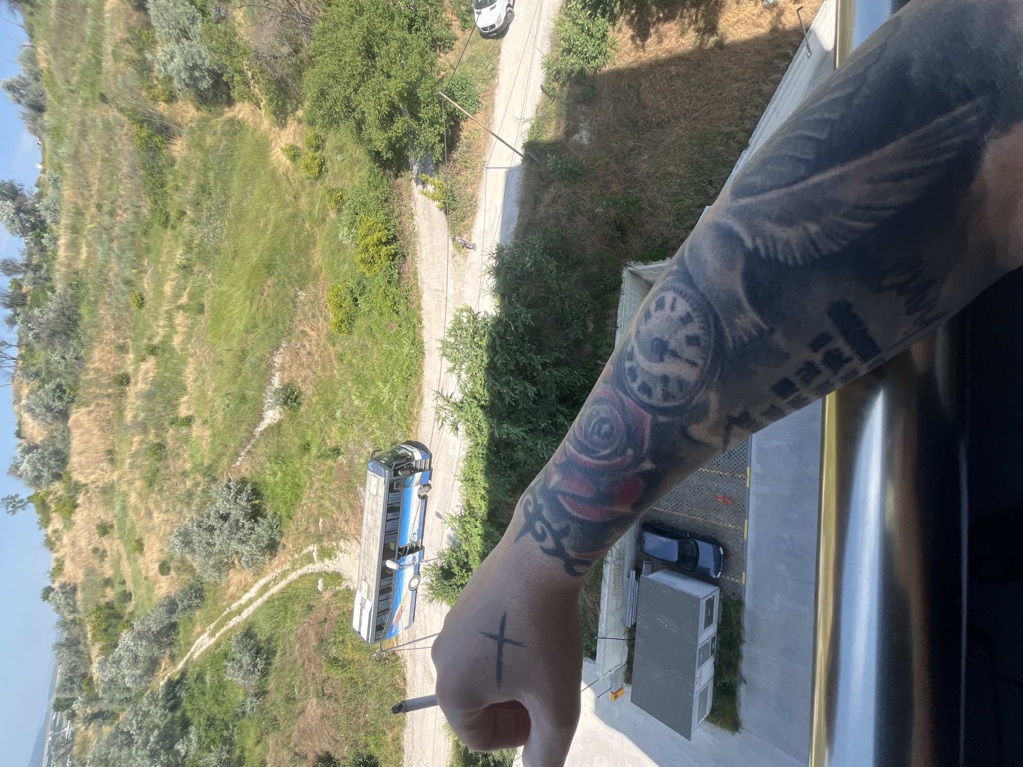 In the city of Varna, Bulgaria, a person's tattooed arm and hand are in the foreground, held up and out of frame from the viewpoint of a balcony. The arm bears a large tattoo of a rose and a clock with wings. The hand has a cross drawn on it. In the background, a blue and white bus is parked on a dirt road. The road winds up a grassy, hilly terrain with scattered trees. A white car is visible further down the road. A small, gray building with a white roof is also present in the lower left corner. The sky is clear and blue.