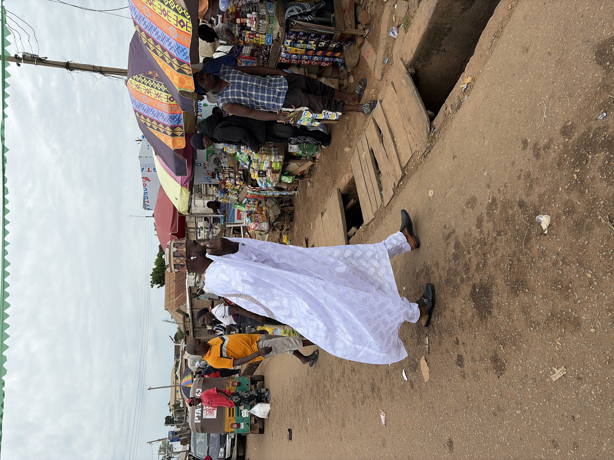 The image depicts a street scene in Gura Top, Nigeria, characterized by a bustling marketplace. A man in a white traditional outfit is walking away from the camera, his face obscured by his hand, seemingly covering his mouth or nose. He is wearing black slip-on shoes. To his left, a man in an orange t-shirt and light-colored shorts walks away from the camera, wearing black flip-flops. Behind him, a person in a red t-shirt and patterned pants stands near a three-wheeled vehicle. In the background, several market stalls are visible, with colorful umbrellas providing shade over displays of goods, including food items, drinks, and other merchandise. A deep drainage ditch runs along the right side of the street, covered in parts by wooden planks. Power lines and poles are visible against an overcast sky. The ground is a mixture of dirt and gravel, with scattered debris.