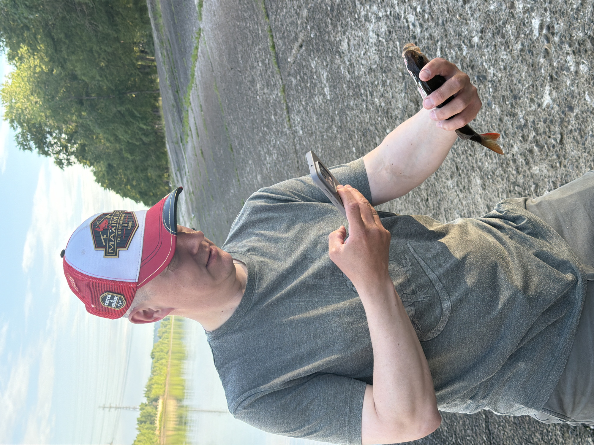 An adult male wearing a gray t-shirt and a red and white trucker hat is standing outdoors next to a body of water. He is holding a small fish in his left hand and a smartphone in his right hand, appearing to take a picture of the fish. The background shows a tree-lined embankment and a hazy sky. The location is the embankment of the Kama River in Chaikovsky, Russia.