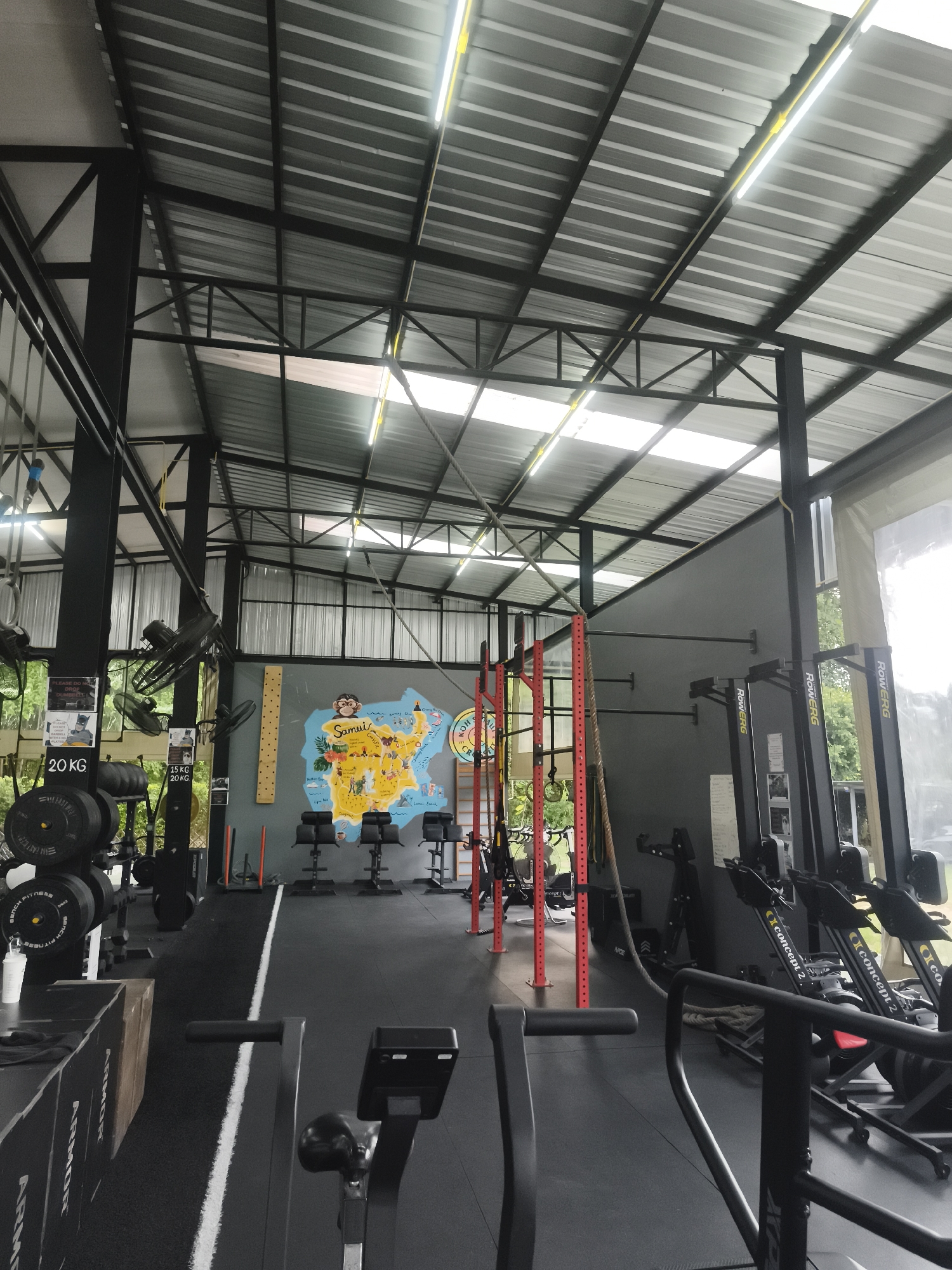 The image captures the interior of a gymnasium, potentially located in Baan Bo Phut, Thailand, given the map of Samui Island on the wall. The space is well-equipped with various exercise machines and equipment, including rowing machines, stationary bikes, weight racks, and a climbing frame. The gym's flooring is black, with a white line marking a lane. Additional equipment such as weight plates and boxes are placed around the weight rack. The walls are painted grey, and the gym is brightly lit, primarily by artificial lights mounted on the metallic ceiling structure. A few ceiling fans provide ventilation. The map of Samui Island on the wall adds a touch of local flavor. Overall, the gymnasium is a functional space designed for diverse workout routines.