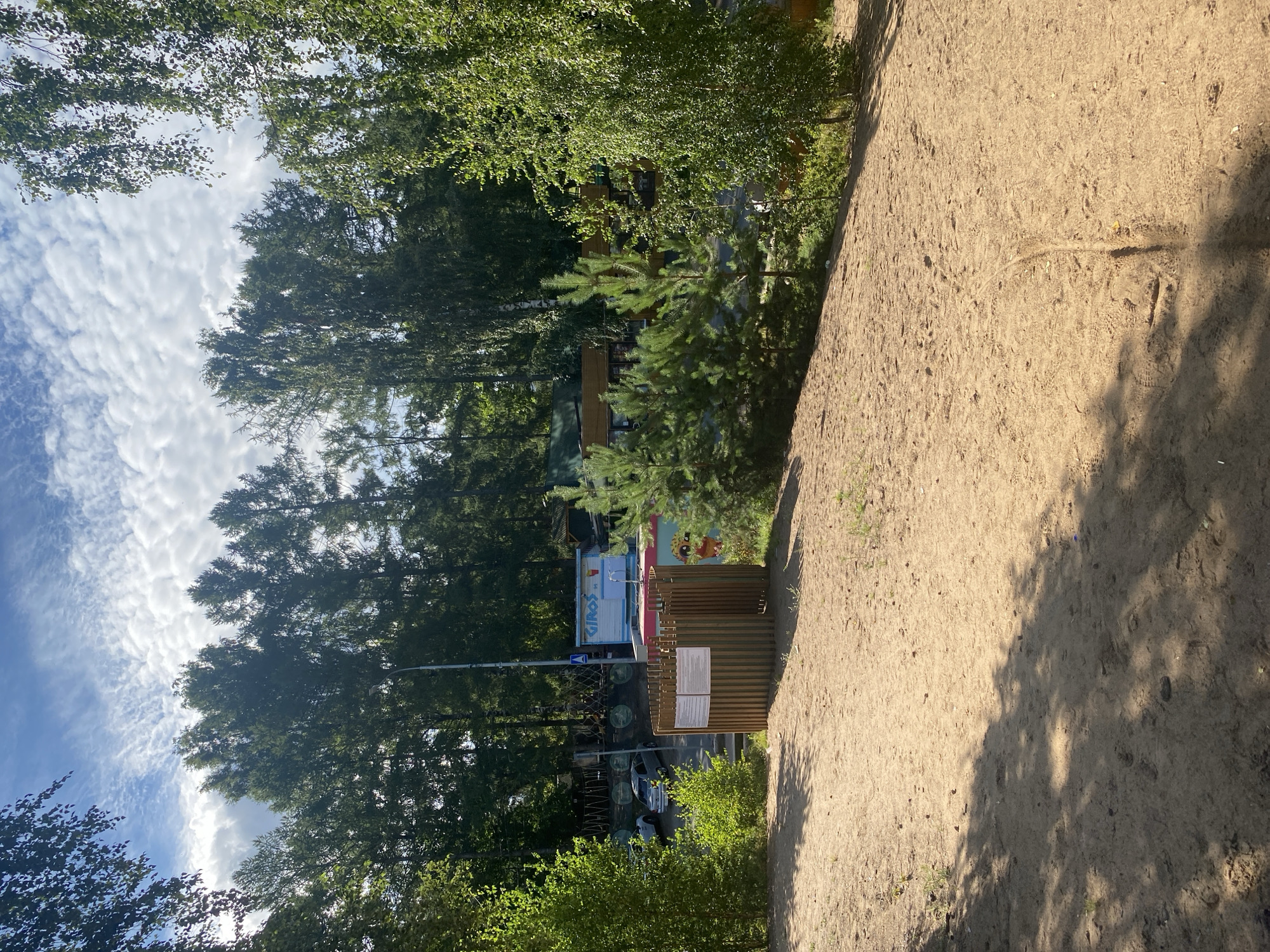 This image depicts a sandy area in what appears to be a park or recreational zone, possibly in the city of Saint Petersburg, Russia. In the foreground, a section of light brown sand is visible, with tire tracks and scattered debris like cigarette butts and small stones. To the left and center, there's a wooden structure, appearing to be a primitive restroom or changing booth, with signs posted on its interior walls. Behind this structure, several cars are parked, and further back, a collection of food stalls or small buildings are situated under a canopy of trees. One of these structures prominently displays the word "GIROS" in blue lettering. The background is dominated by a dense forest of tall trees, reaching towards a sky filled with white, wispy clouds against a blue backdrop. The overall impression is that of an outdoor public space, possibly a beach or a wooded area with amenities.