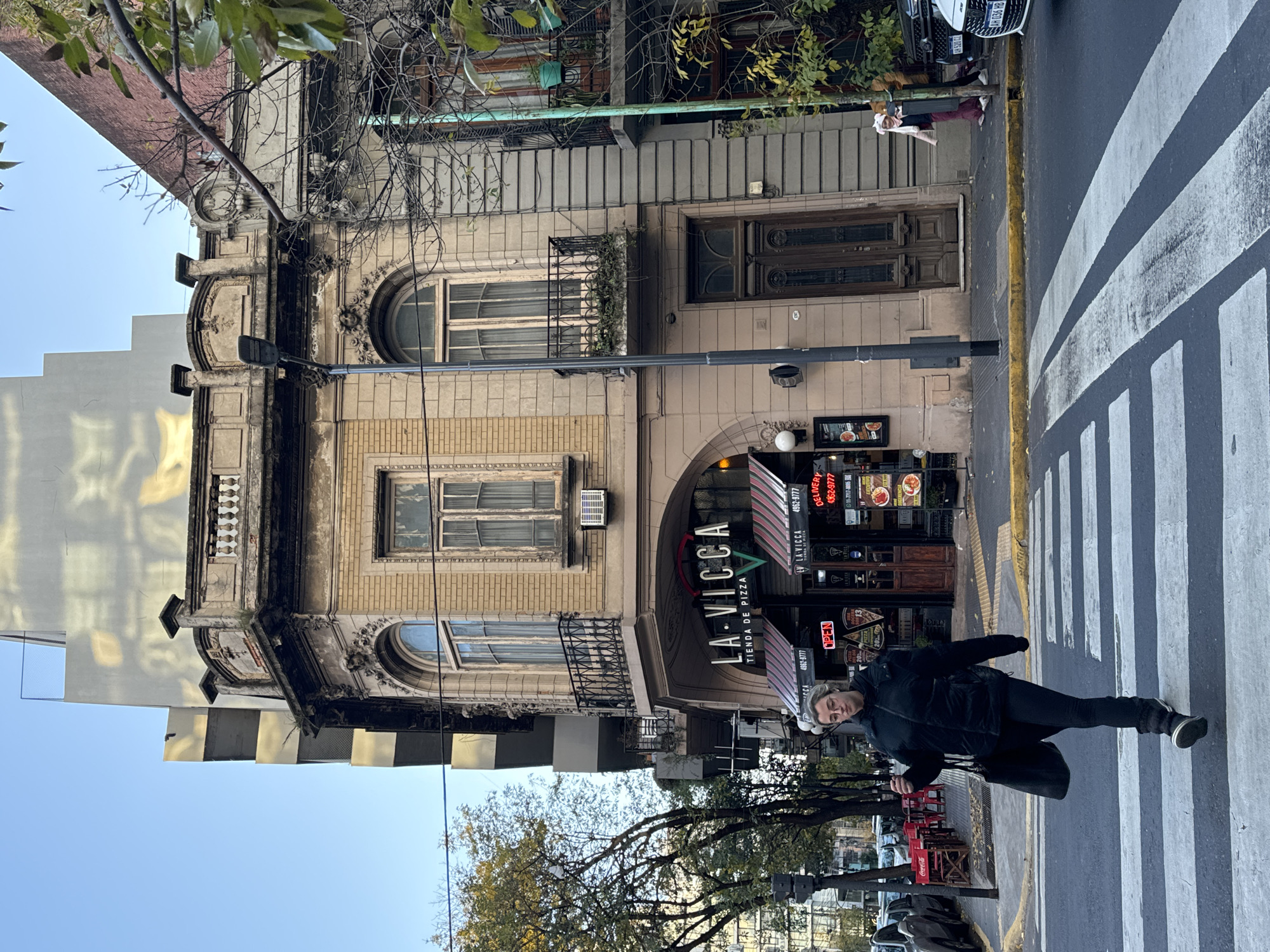 The image captures a street scene in Buenos Aires, Argentina. A woman dressed in black is mid-stride in a crosswalk marked with broad white stripes, appearing to be walking diagonally across the street. Behind her is a restaurant named "La Vicca Tienda de Pizza," identified by its sign and banners displaying phone numbers and pizza imagery. A tree, partially obscuring the foreground, suggests a tree-lined street. Behind the restaurant is an older building with architectural details, including arched windows and decorative elements. A gray high-rise building is reflected in the building across the street. Parked cars are visible along the street, including a car with a license plate that has the letters "HB" on it. Also, red chairs and tables are seen in front of the restaurant. The sky is partly clear, suggesting either early morning or late afternoon.