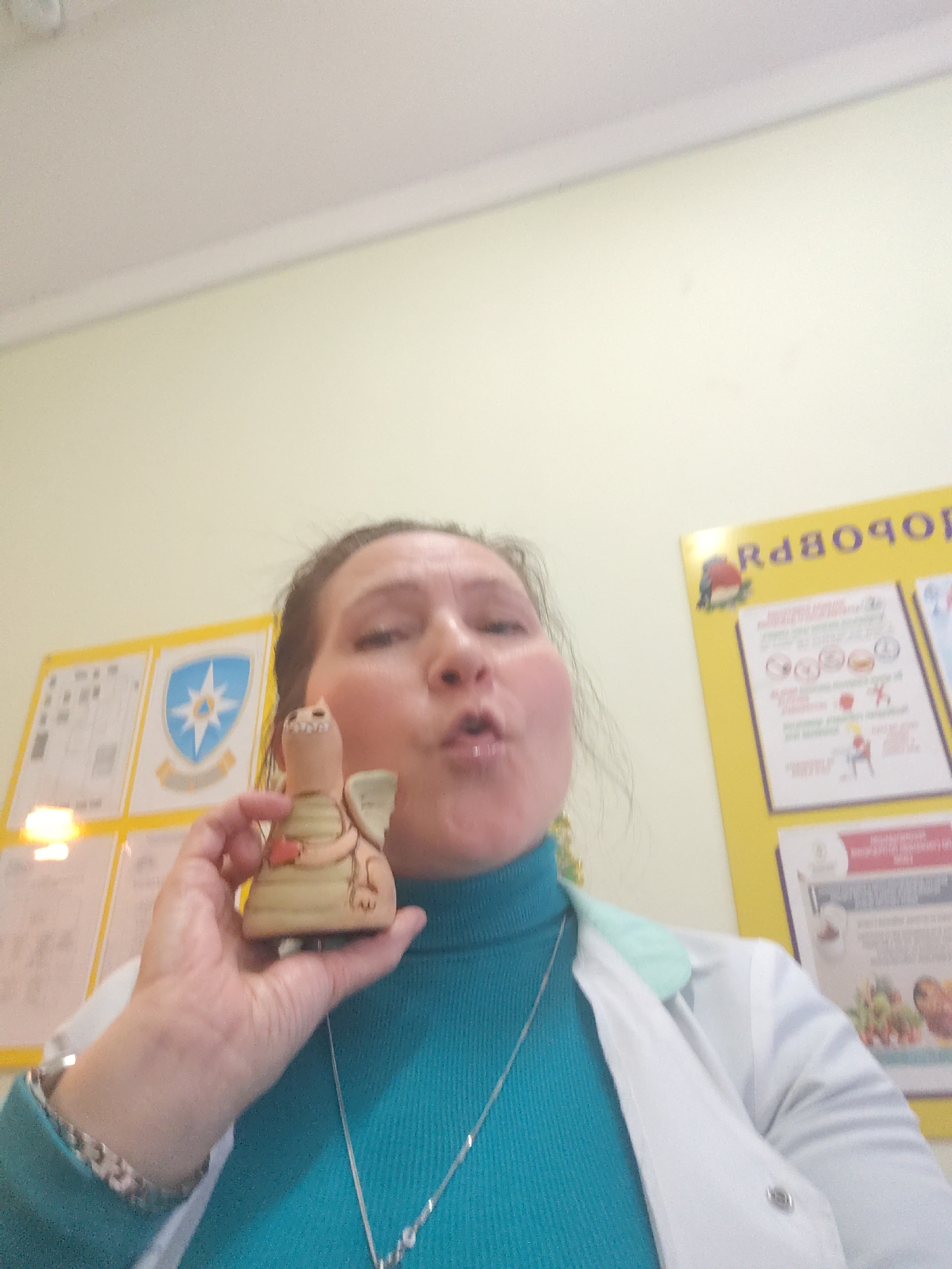 A woman holds a ceramic dragon figurine. She is wearing a turquoise turtleneck and a white lab coat, along with jewelry including a silver chain and bracelet. She stands in a room with yellow walls, where posters are visible. One poster displays the text "ЗДОРОВЬЕ". The image was likely taken in Sarapul, Russia.
