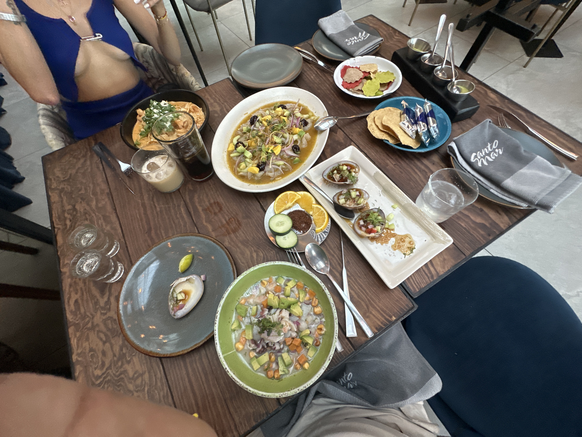 The scene depicts a wooden table in a restaurant, laden with an assortment of dishes. The dining environment is well-lit, indicating daytime. There are plates filled with ceviche, tostadas, and tortilla chips. The ceviche is served in various forms: in a large oval dish, in a green bowl, and in oyster shells on a white rectangular plate. A sauce sampler is present on the table along with water and other drinks. Silverware and napkins are neatly arranged. A female diner is seated at the table, wearing a blue top. The restaurant appears to be in the city of Santiago de Querétaro, Mexico, as indicated by the "Santo Mar" napkin which is a restaurant located there.