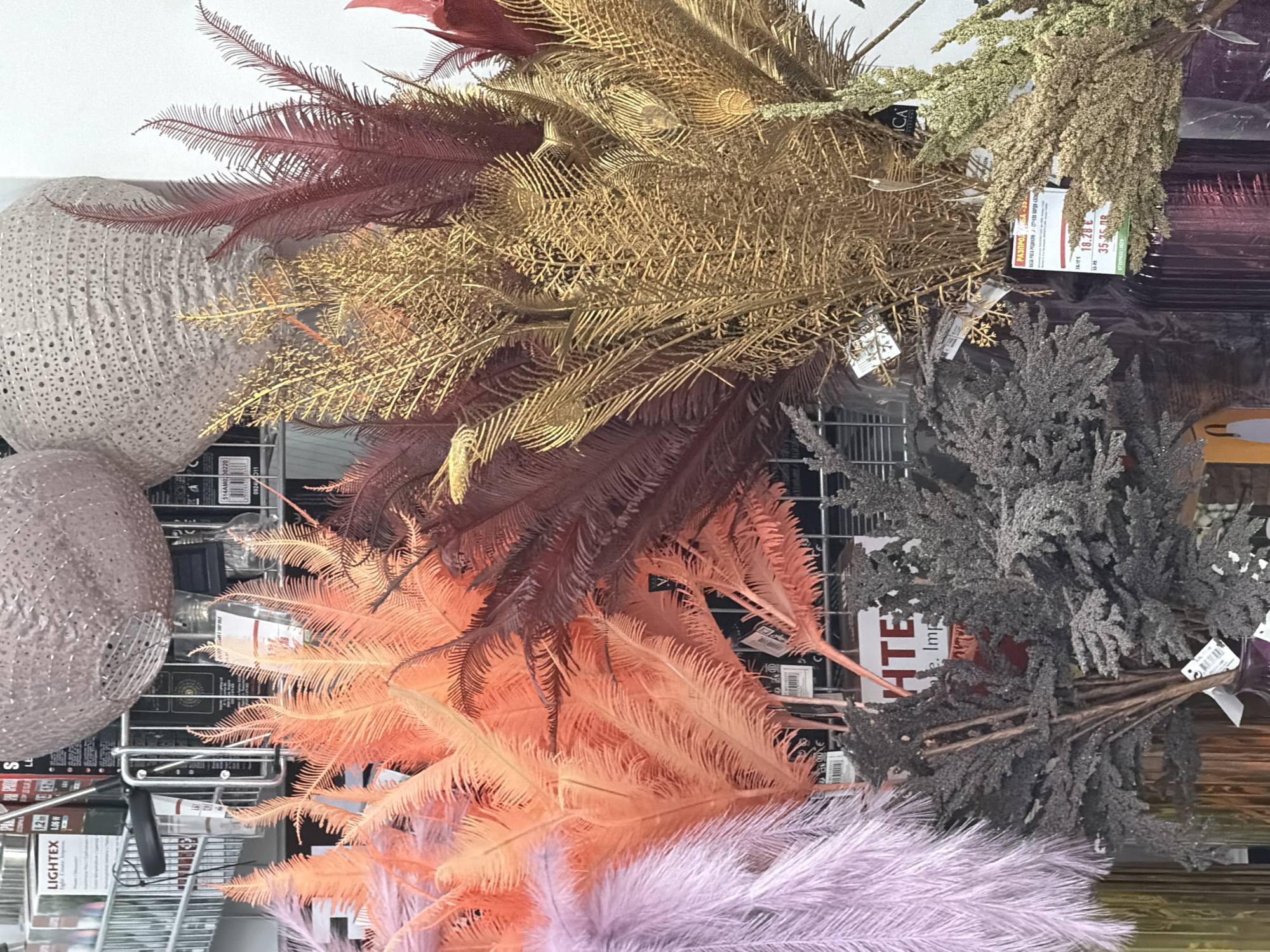 The image shows a close-up, eye-level, medium shot of an assortment of artificial floral decorations, likely displayed for sale in a retail environment. The primary subjects are several large, feathery branches in various vibrant colors, including a pale lavender, a bright coral orange, and a deep reddish-brown. Interspersed with these are branches with a metallic gold finish, resembling dried grasses or palm fronds. In the foreground, to the left and center, are bunches of darker, more subdued artificial foliage, possibly covered in a glittery material, appearing grey or dark gray.
Behind the floral arrangements, a metal wire rack is visible, suggesting a display stand. Product packaging and labels are partially discernible on the rack, with some text in what appears to be Cyrillic script, indicating a location in Bulgaria. One label clearly displays the currency symbol "€" and prices such as "10.28 €" and "35.75 BGN", with "BGN" being the Bulgarian Lev. Another visible label reads "LIGHTEX" along with wattage information, likely for lighting products. Two large, decorative, textured spheres, possibly lampshades or decorative orbs, are positioned at the top of the frame, one appearing textured and beige, the other a mottled pinkish-brown.
The overall scene is brightly lit, with the focus on the textures and colors of the artificial plants. The image composition is such that the viewer's eye is drawn to the vibrant and varied textures of the decorative branches. There are no people or active interactions depicted. The environment appears to be an indoor retail space, such as a home goods store or a craft supply store, possibly within the city of Plovdiv, Bulgaria, given the textual clues on the product labels.