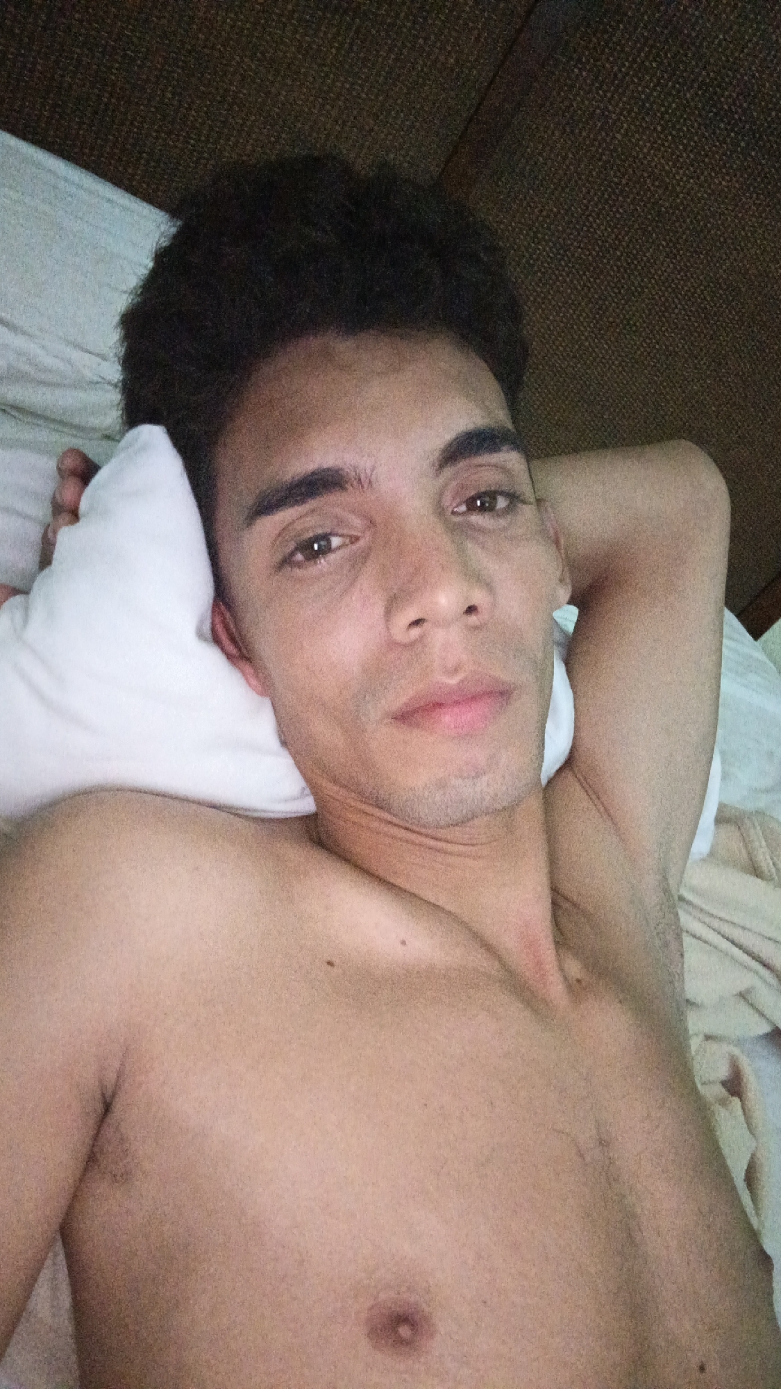 A shirtless man is lying on a bed. He is propped up on a white pillow with his left arm under his head and his right arm behind his head. He has dark, curly hair, brown eyes, and a neutral expression. The background consists of a light-colored sheet and a dark brown headboard. It is not possible to identify the location as Playa El Agua, Venezuela, from the image alone.
