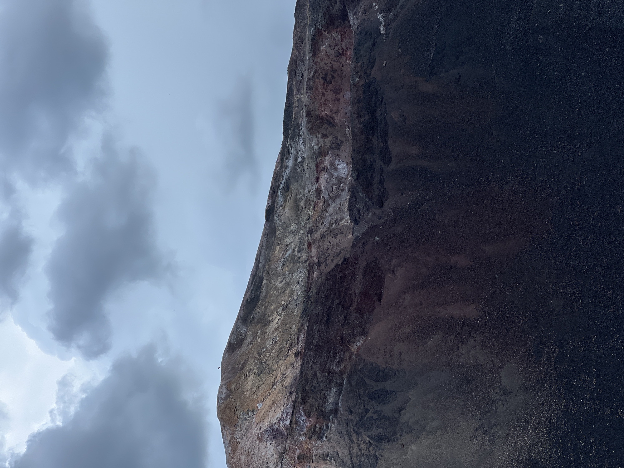 An image captures a volcanic landscape under a cloudy sky. The foreground is dominated by dark, rough volcanic rock, which transitions into a lighter, more varied slope of reddish and yellowish hues further up the mountain. A small, dark figure, potentially a person or an animal, is visible standing on the crest of the volcano. The sky is filled with heavy, gray clouds, suggesting overcast weather. There is no discernible urban environment or indication of the city of Unknown, Nicaragua within the frame.