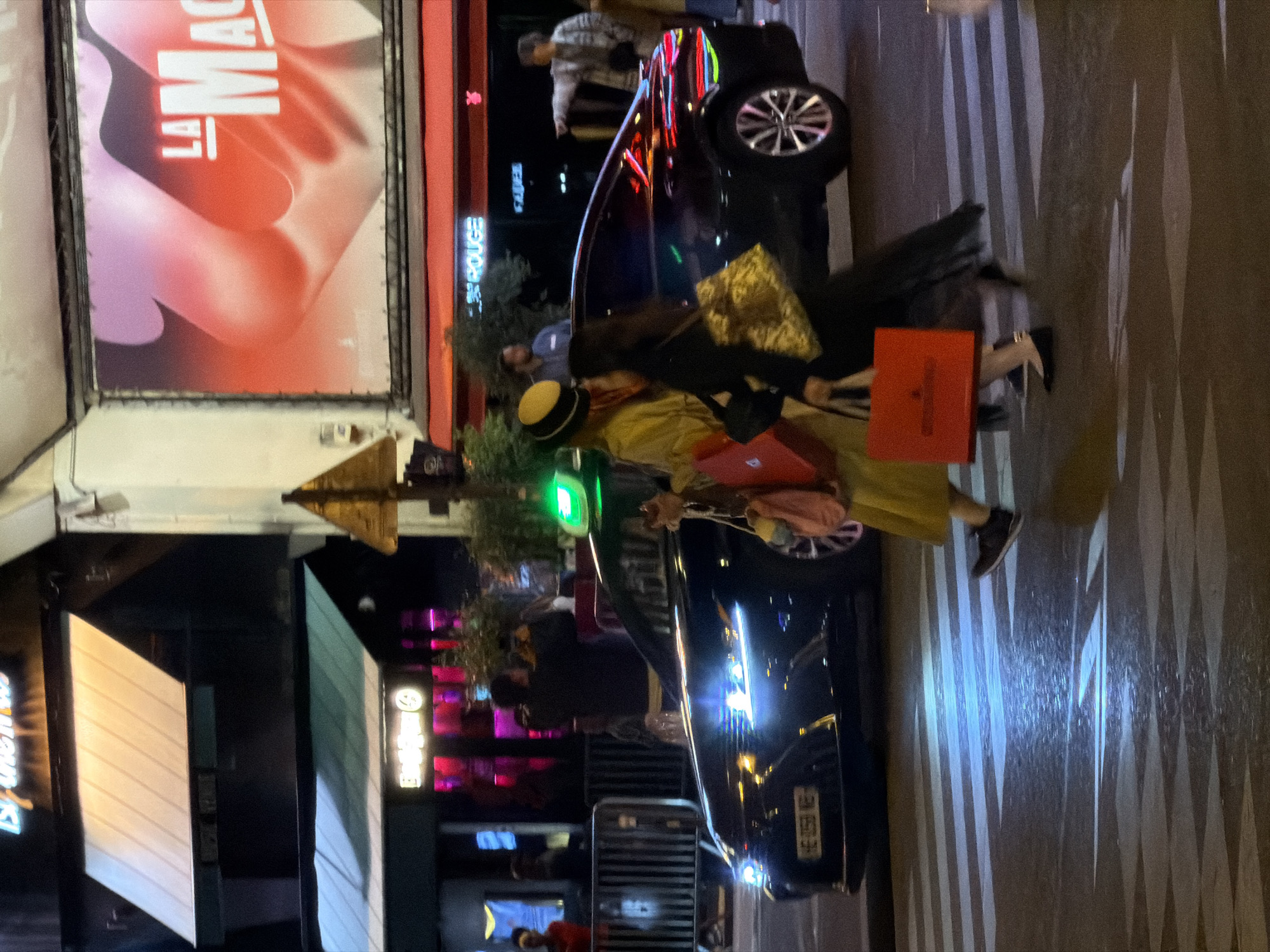The image depicts a nighttime street scene in Paris, France, focusing on a pedestrian crossing the street near the Moulin Rouge cabaret, identifiable by its iconic signage. A black car is stopped at a crosswalk, its headlights illuminating the patterned street surface. A person wearing a yellow coat and a hat is crossing in front of the car, carrying shopping bags, including a prominent red one. In the background, neon signs and lights from surrounding businesses illuminate the street. Pedestrians can be seen in the background near the vehicle. The overall scene captures the vibrant atmosphere of the Parisian nightlife.