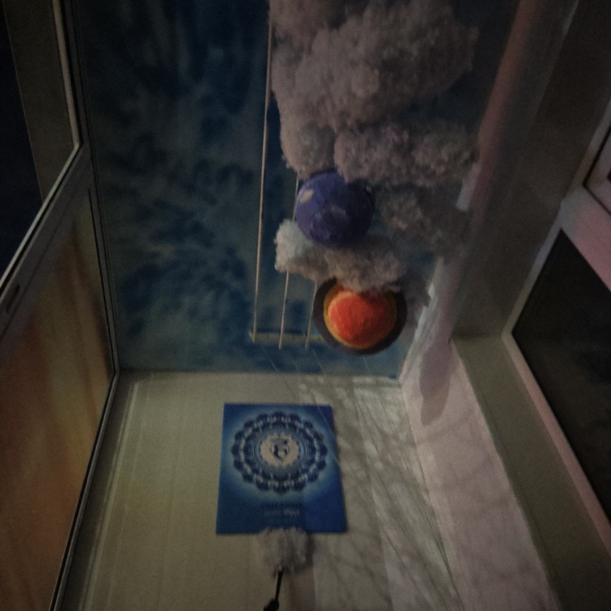 A low-angle interior shot reveals a room with multiple design elements. The ceiling is painted to resemble a blue sky with white clouds, and three-dimensional representations of planets, surrounded by cotton-like material, hang from a metal rack. A blue square poster, printed with a mandala-like design, is mounted on a wall; a white duster is placed near it. Light from the window illuminates the scene, casting shadows on the wall. A framed dark rectangle hangs on the adjacent wall. The location is likely in Rostov, Russia.