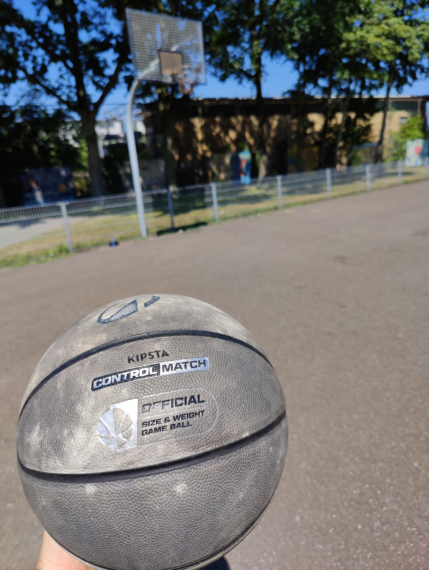 A grey basketball with the Kipsta logo and "CONTROL MATCH OFFICIAL SIZE & WEIGHT GAME BALL" printed on it is held in a person's hand. The basketball is in the foreground, and the background shows a basketball court with a hoop and net, trees, and a building. The scene appears to be outdoors on a sunny day. The location is the city of Breda, Netherlands.