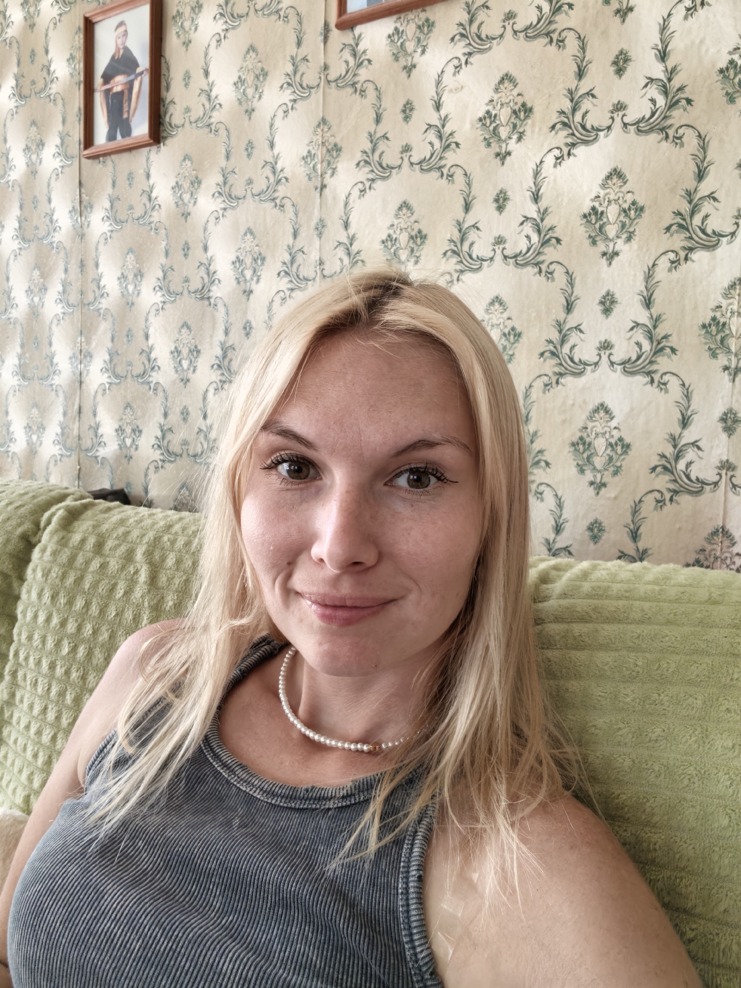 A young, fair-skinned woman with blonde hair is taking a selfie while sitting on a textured green sofa. She is wearing a grey, ribbed tank top and a pearl necklace. The background consists of a wall covered in ornate, light-colored wallpaper with a repeating pattern of green floral designs. To the left of the woman, a framed picture hangs on the wall, showing a person in a black outfit. The setting appears to be indoors, possibly a living room. The image does not contain information about the city of Sterlitamak, Russia.