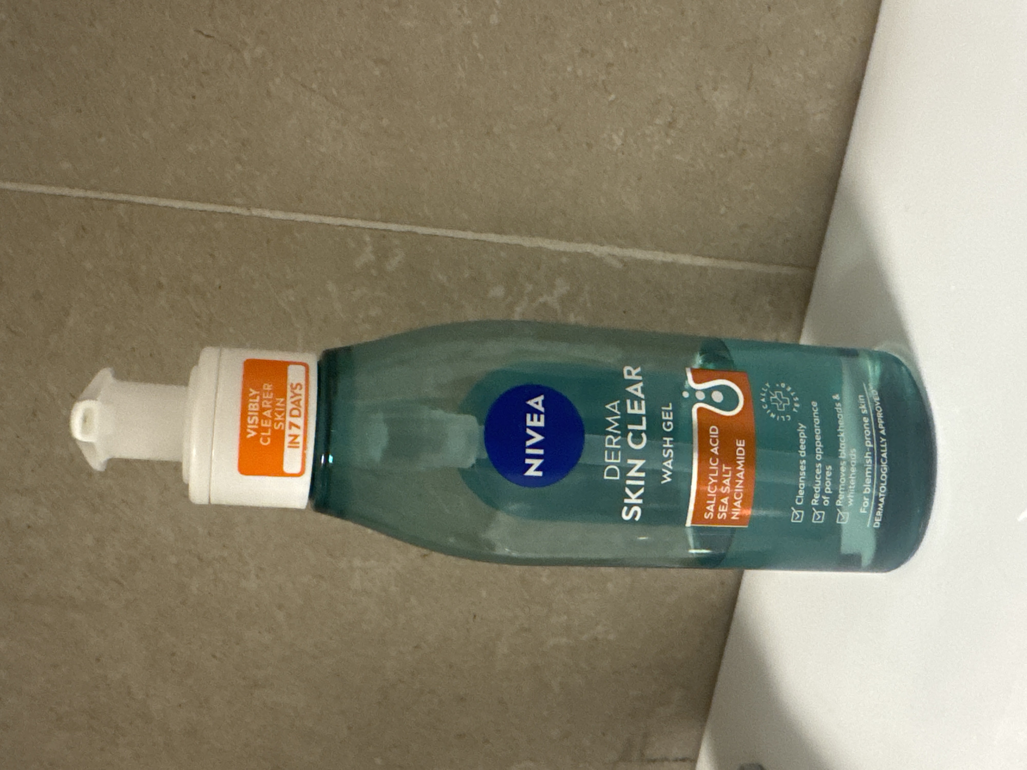 A tall, teal-colored plastic bottle of Nivea Derma Skin Clear Wash Gel stands upright on a white surface, possibly a countertop, in front of a beige tiled wall. The bottle has a white pump dispenser at the top, with a translucent white plastic cap covering the opening. A prominent orange and white label on the upper part of the bottle reads "VISIBLY CLEARER SKIN IN 7 DAYS". The main body of the bottle features the Nivea logo in a blue circle, followed by "DERMA SKIN CLEAR WASH GEL". Below this, a rectangular label displays "SALICYLIC ACID SEA SALT NIACINAMIDE" in white lettering. A smaller graphic of two bubbly circles is also visible on this label. The lower portion of the bottle lists benefits with checkmarks: "Cleanses deeply", "Reduces appearance of pores", "Removes blackheads & whiteheads", and "For blemish-prone skin". The text "DERMATOLOGICALLY APPROVED" is at the very bottom. The background is a beige tiled wall with visible grout lines. The lighting is even, suggesting an indoor setting. There are no people or animals present in the image.