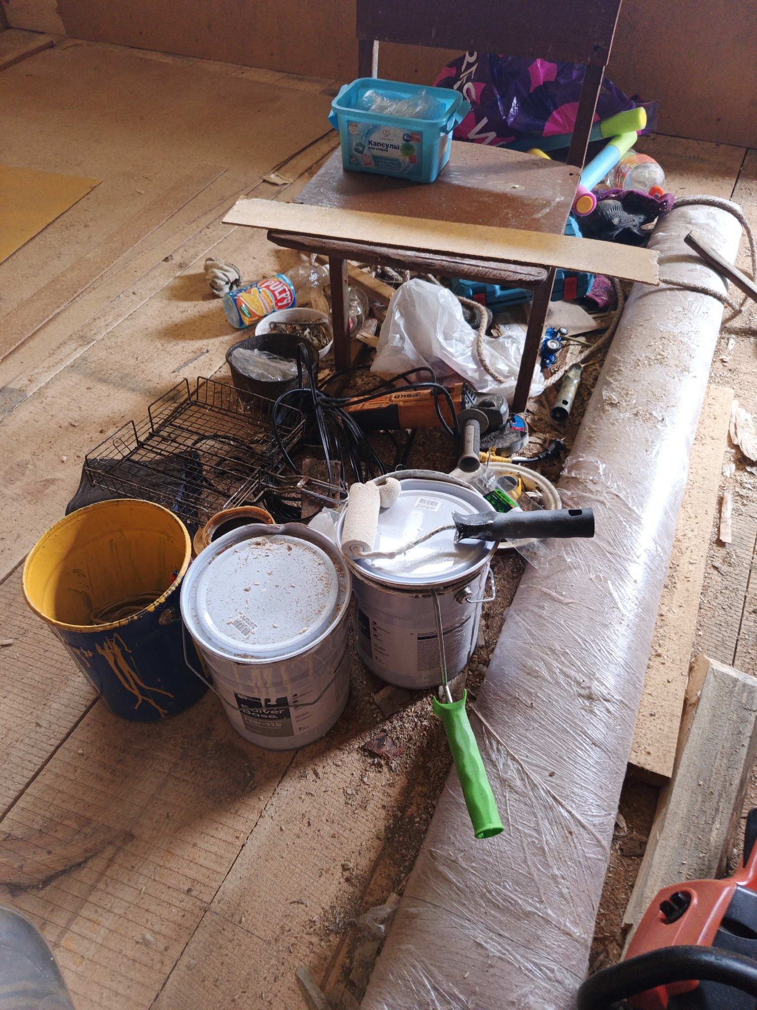 The image depicts a cluttered room with wooden floors and walls, likely in a storage or workshop area in Islambaevo, Russia. The floor is covered in sawdust and debris. In the foreground, there are two large paint buckets, one blue and yellow and another white, with a paint roller resting on top of the white bucket. Next to the paint buckets is a yellow bucket and a black wire rack. Further back, a wooden table is laden with various items, including a blue plastic container with what appears to be cleaning supplies, a rolled-up mat or insulation wrapped in plastic, and a bundle of colorful pool noodles. An orange power tool, possibly a grinder, is visible in the lower right corner of the frame. The overall impression is one of disarray and ongoing or recently completed construction or renovation work.