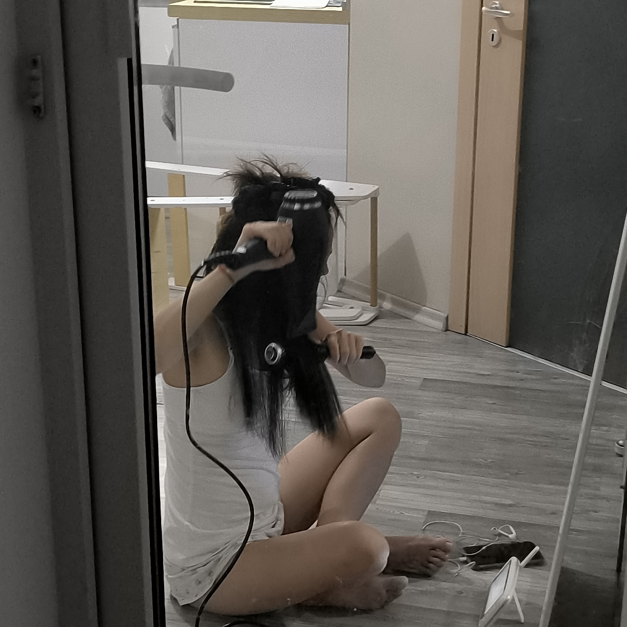 In Varna, Bulgaria, a young woman sits on the floor of a room, likely a bedroom or living area, styling her long dark hair. She is wearing a white tank top and shorts. She is holding a black hairdryer and a round brush, actively using them to straighten her hair. The floor is covered with grey laminate flooring. In the background, there is a light-colored shelf unit and a doorway with a light-colored doorframe and a dark door. A partial view of a mirror is visible on the right side of the image, reflecting some of the room and the woman. Various objects are scattered on the floor near her, including a smartphone, headphones, and a small white stand. The scene appears to be captured through a reflective surface, possibly a window or a glass door, as evidenced by the dark frame on the left.