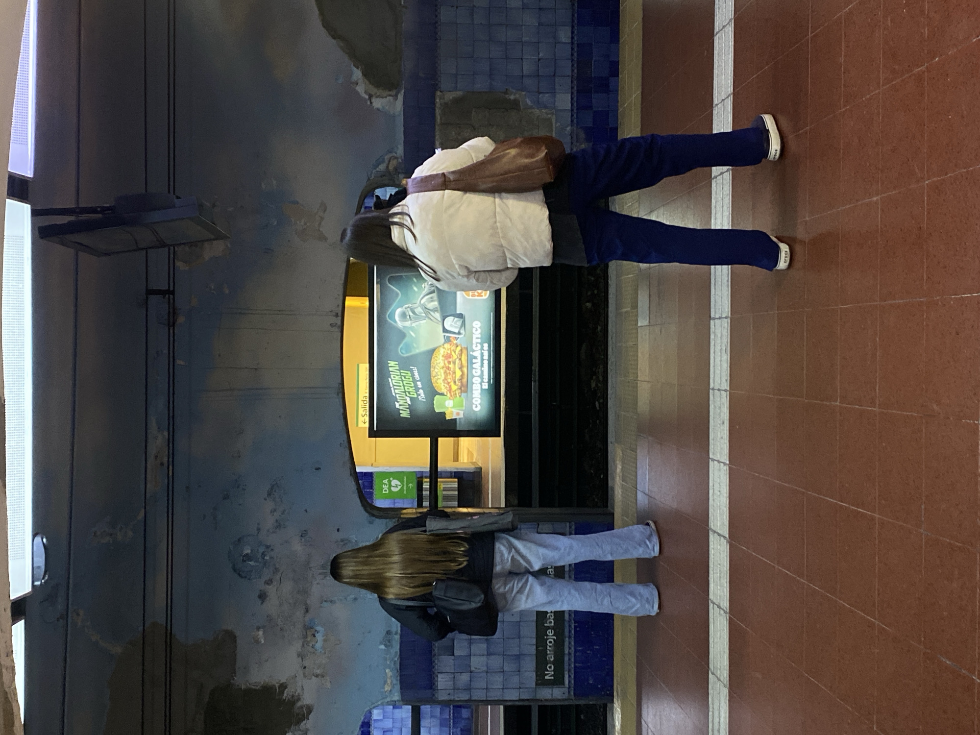Here's a description of the image:
The photograph depicts a subway platform in Buenos Aires, Argentina. Two women stand facing an advertisement for "The Mandalorian & Grogu" movie and a McDonald's "Combo Galactico" meal.
One woman is wearing a white puffer jacket, blue pants, and has a brown shoulder bag. The second woman wears a dark jacket and light-colored jeans.
The platform's design includes blue and white tiled walls, a yellow edge, and a red-tiled floor. A sign pointing towards an exit ("Salida") is visible. A warning against littering ("No arroje basura") is printed on the platform edge. A rectangular light fixture hangs above.