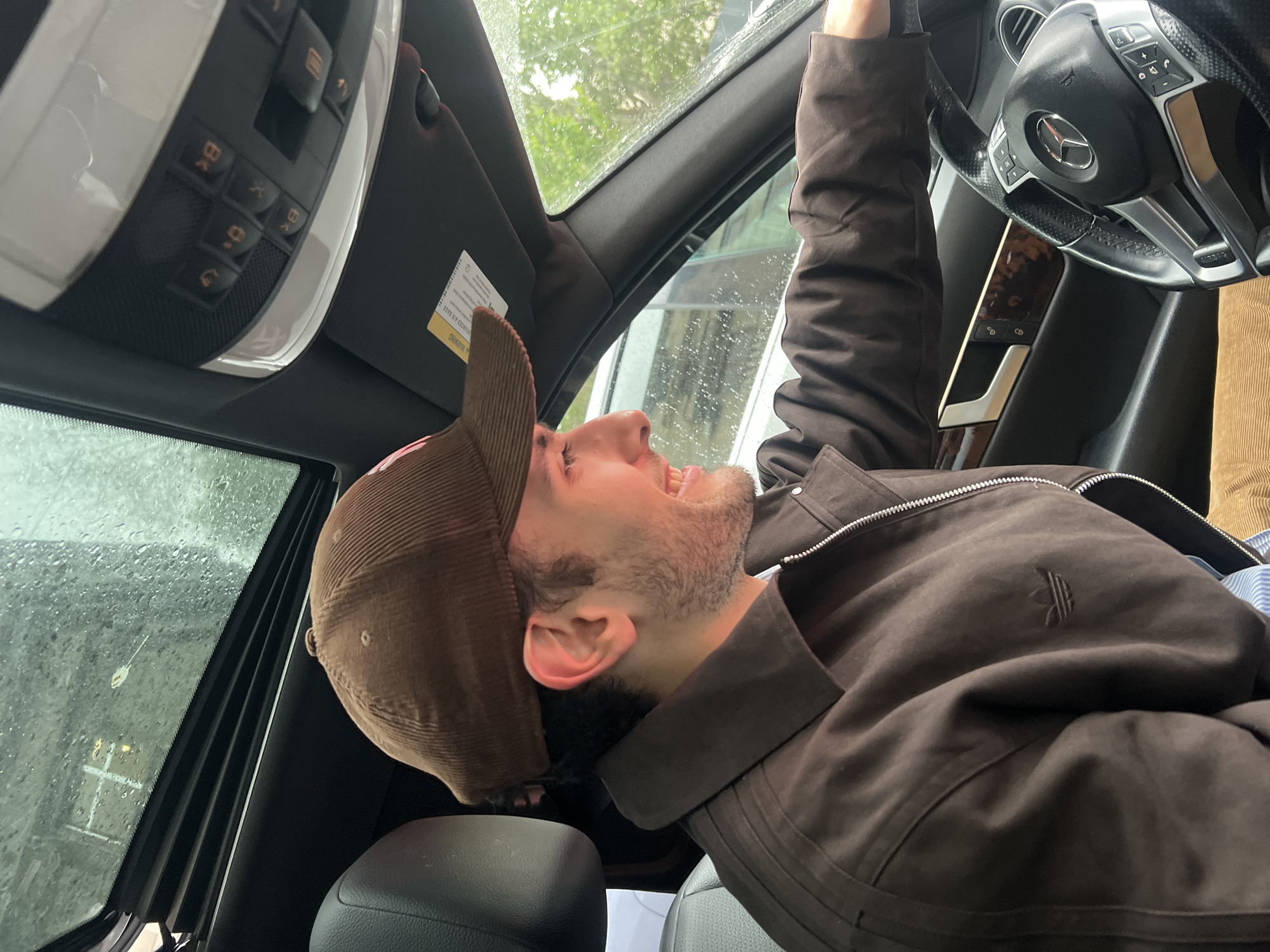A man wearing a brown corduroy baseball cap and a brown Adidas jacket is inside a Mercedes-Benz. He is in the driver's seat, with his right hand on the steering wheel and his head tilted back. The sunroof is open, and raindrops are visible on the window. The interior of the car has a black and brown color scheme. The location cannot be determined as Yerevan, Armenia, from the visual data provided.