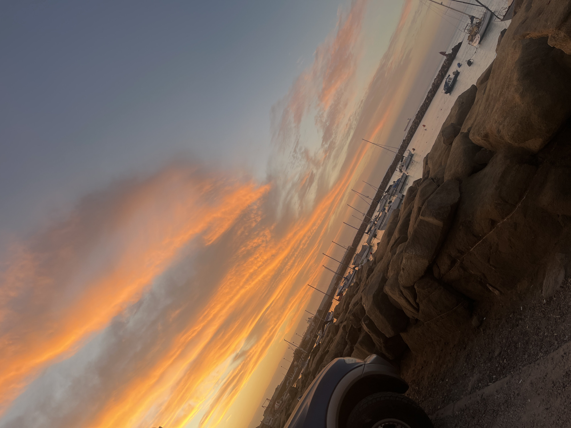 The image depicts a sunset scene in Arica, Chile. The sky is filled with orange and grey clouds. In the distance, there's a harbor area with boats and a breakwater. The right side of the image is dominated by a rocky outcrop. The wheel well of a car is visible in the lower center of the image.
