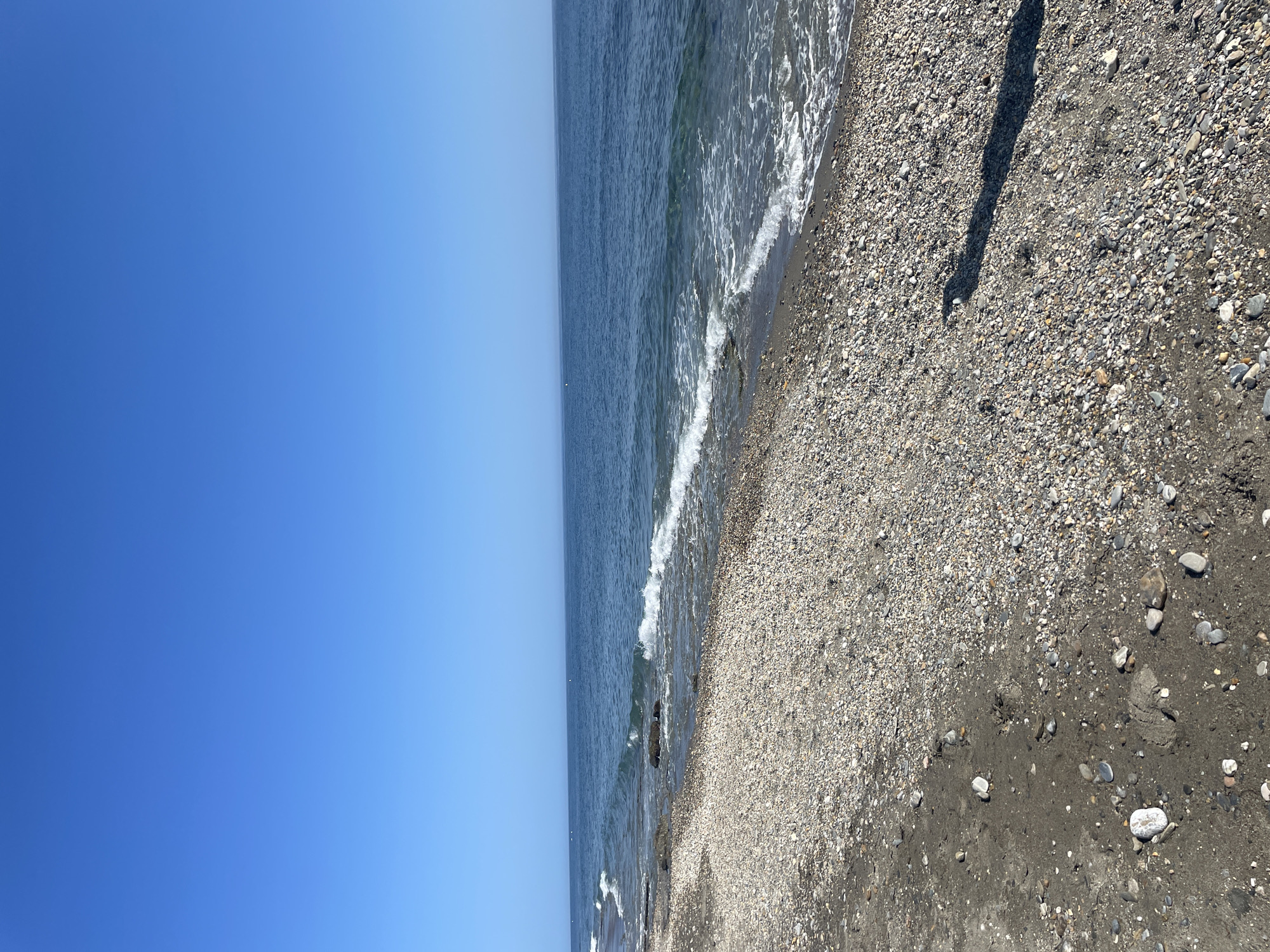 The image depicts a serene beach scene under a clear, vibrant blue sky. The foreground is a pebble-strewn beach with a mixture of dark sand and a multitude of small, multicolored pebbles. Some larger rocks are visible, and a shadow of an unseen object stretches across the beach from the right side of the frame. The middle ground features the gentle, foamy waves of a blue sea washing onto the shore. In the distance, the sea merges seamlessly with the horizon, where a faint, hazy outline of land can be discerned. The overall impression is one of a peaceful, sunny day at the coast. The location is identified as a beach in Unknown, Spain.