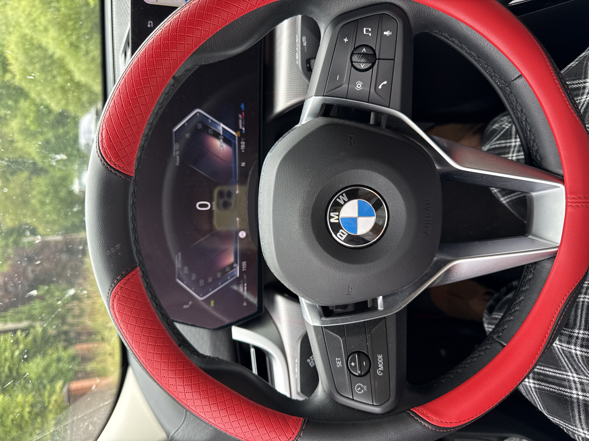The image shows a close-up view of a BMW steering wheel with a red and black cover, inside a car. The car's digital dashboard is visible, displaying speed, temperature, and other information. The steering wheel has various buttons for controlling functions like cruise control, audio, and phone calls. The driver's checkered pants are visible below the steering wheel, indicating someone is driving. The scene appears to be in an outdoor environment, possibly on a street, given the blurred greenery and trees visible through the windshield. The specific location of Russko-Vysotskoye Sel'skoye Poseleniye, Russia cannot be determined from the visual information present in the image.