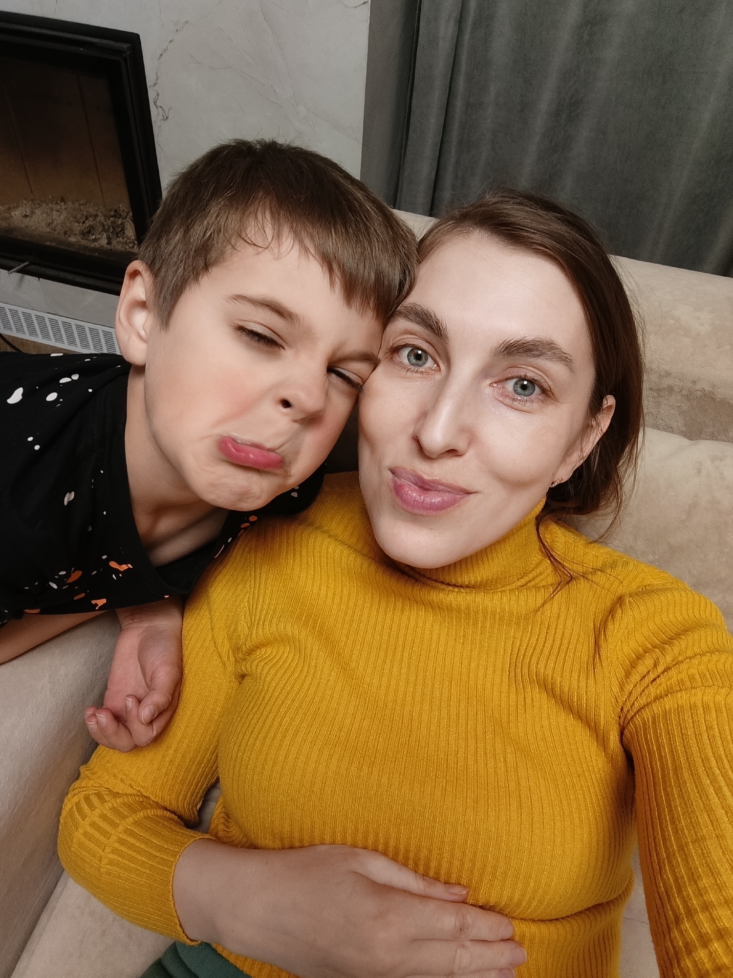 A selfie features a woman and a child. The woman, on the right, wears a mustard yellow turtleneck sweater and has light skin, brown hair, and blue eyes. She is smiling slightly at the camera. The child, on the left, has short brown hair and is making a comical face with pursed lips and one eye squinted shut. The child is wearing a black shirt with white and orange paint splatter designs. The child's hand rests on the woman's shoulder. They are sitting on a light-colored upholstered sofa.
In the background, on the left, is a dark fireplace insert. To the right of the fireplace, the wall appears to have a marble-like texture. A dark grey curtain is visible on the right side of the image.
The image was taken in an interior setting; the precise location within the city of Chaikovsky, Russia, cannot be determined from the provided image.