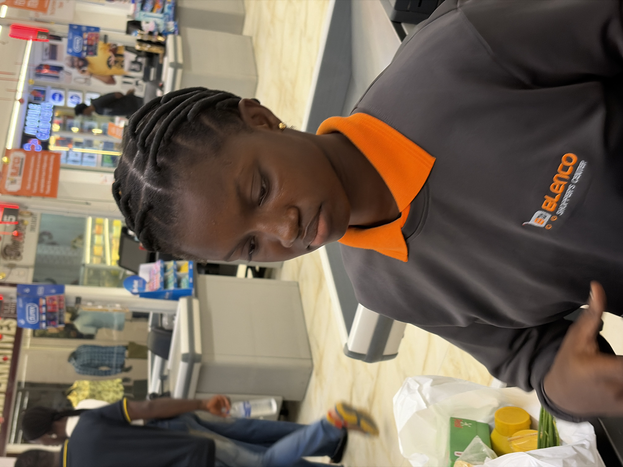 The image shows a cashier working at the "Elenco Shoppers Center" checkout in Igboefon, Nigeria. She has dark skin, braided hair, and is wearing a grey shirt with an orange collar, which bears the logo of the company. A bag with items is on the counter, including yellow containers and green vegetables. In the background, other people can be seen in the store, as well as shelves stocked with various items.